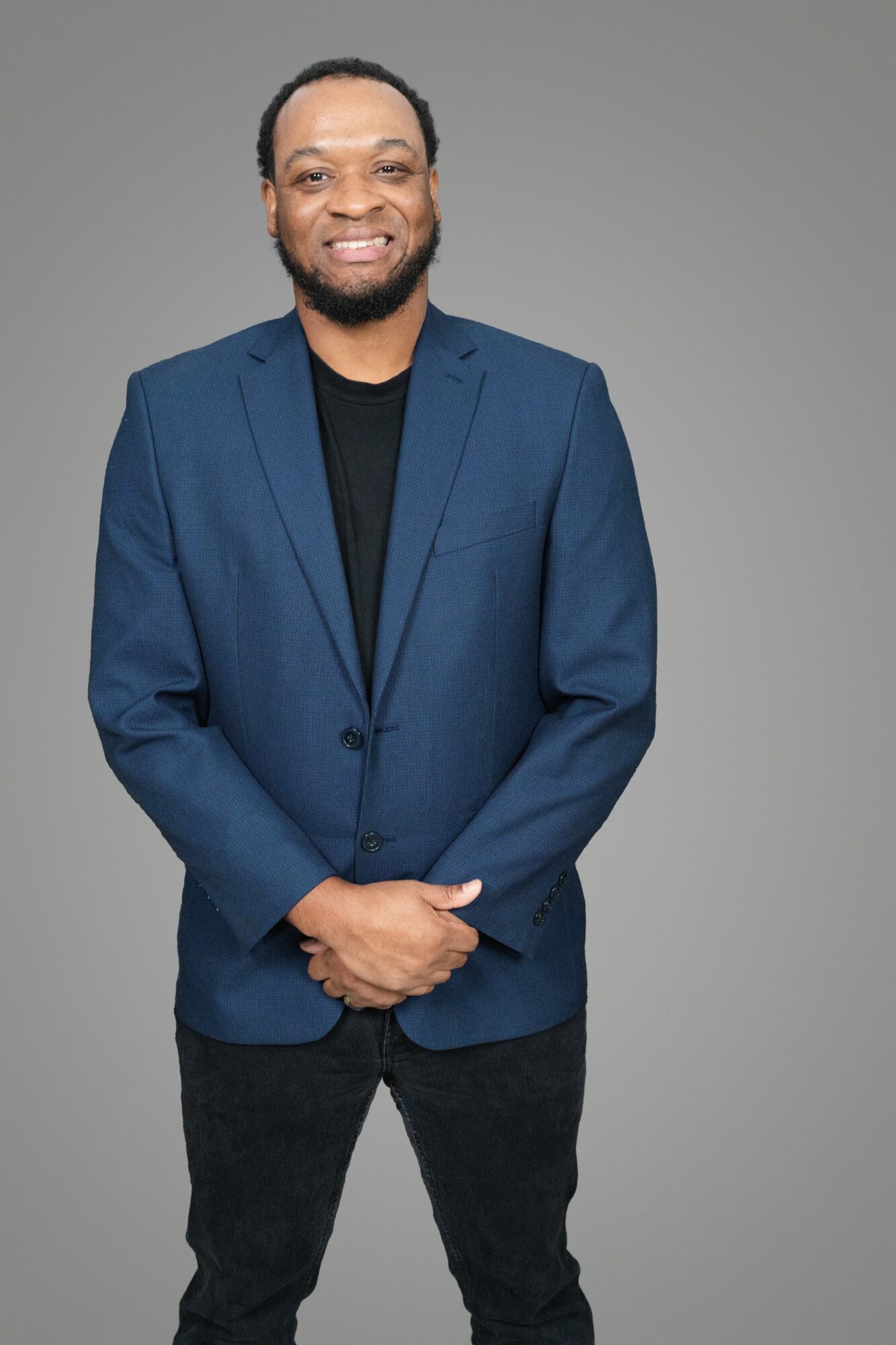 Man with a beard in a blue blazer and black shirt, smiling, standing against a gray background.