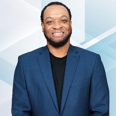 Man with short curly hair and beard wearing a dark blue suit and black shirt, smiling, against a light geometric background.