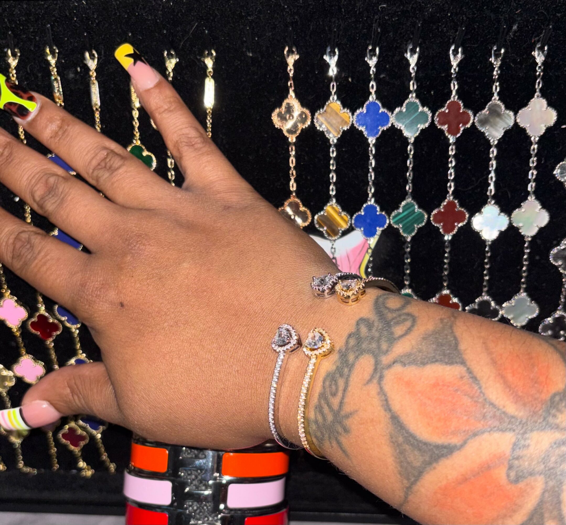 Hand with jewelry and colorful nail polish, tattooed arm, jewelry display in background, black backdrop.