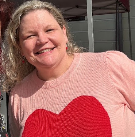 Smiling woman with curly hair wearing a pink shirt with a red heart design outdoors.