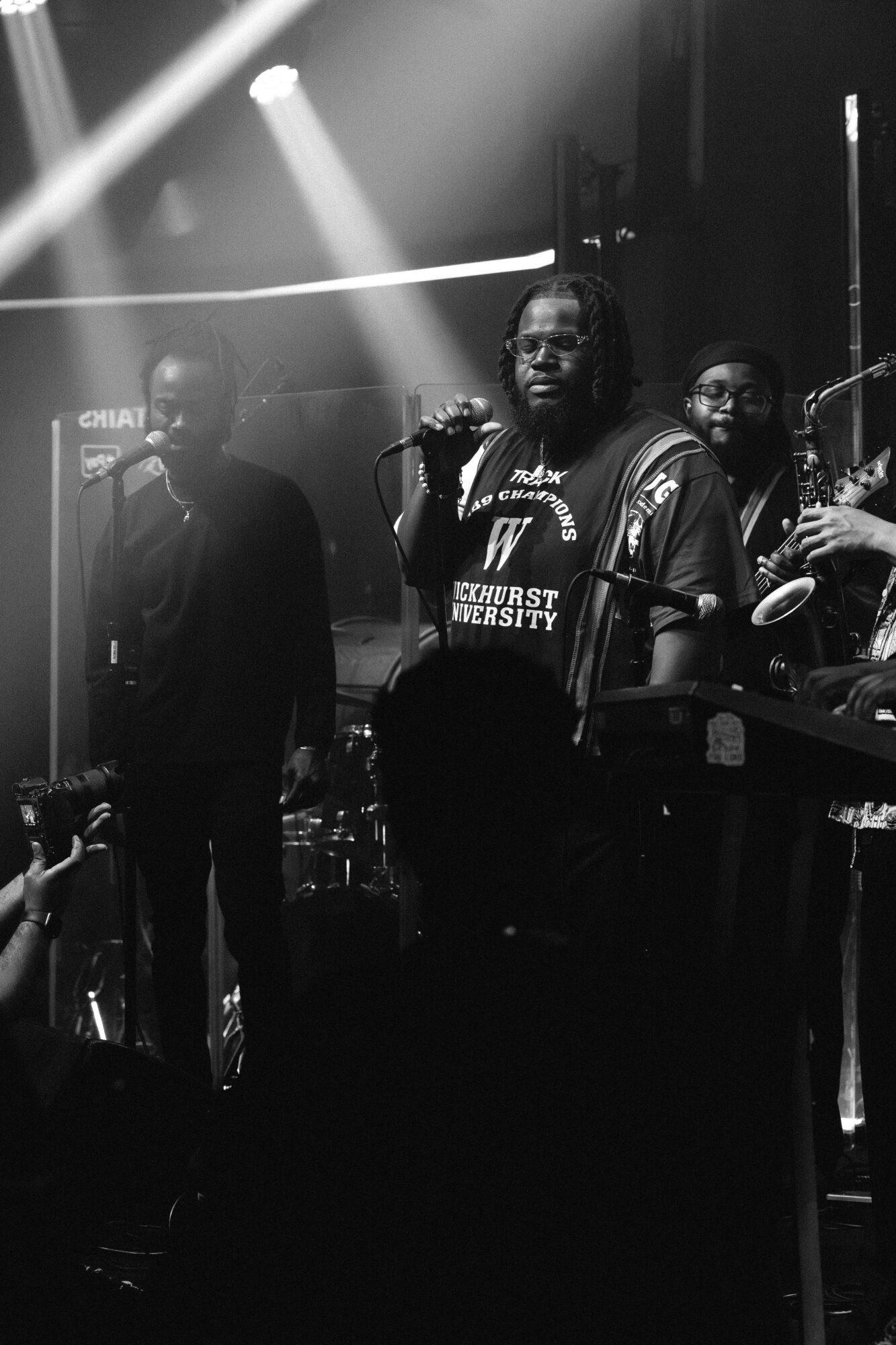 Three musicians perform on stage with lighting, one holding a microphone, others playing instruments, in a dark setting.