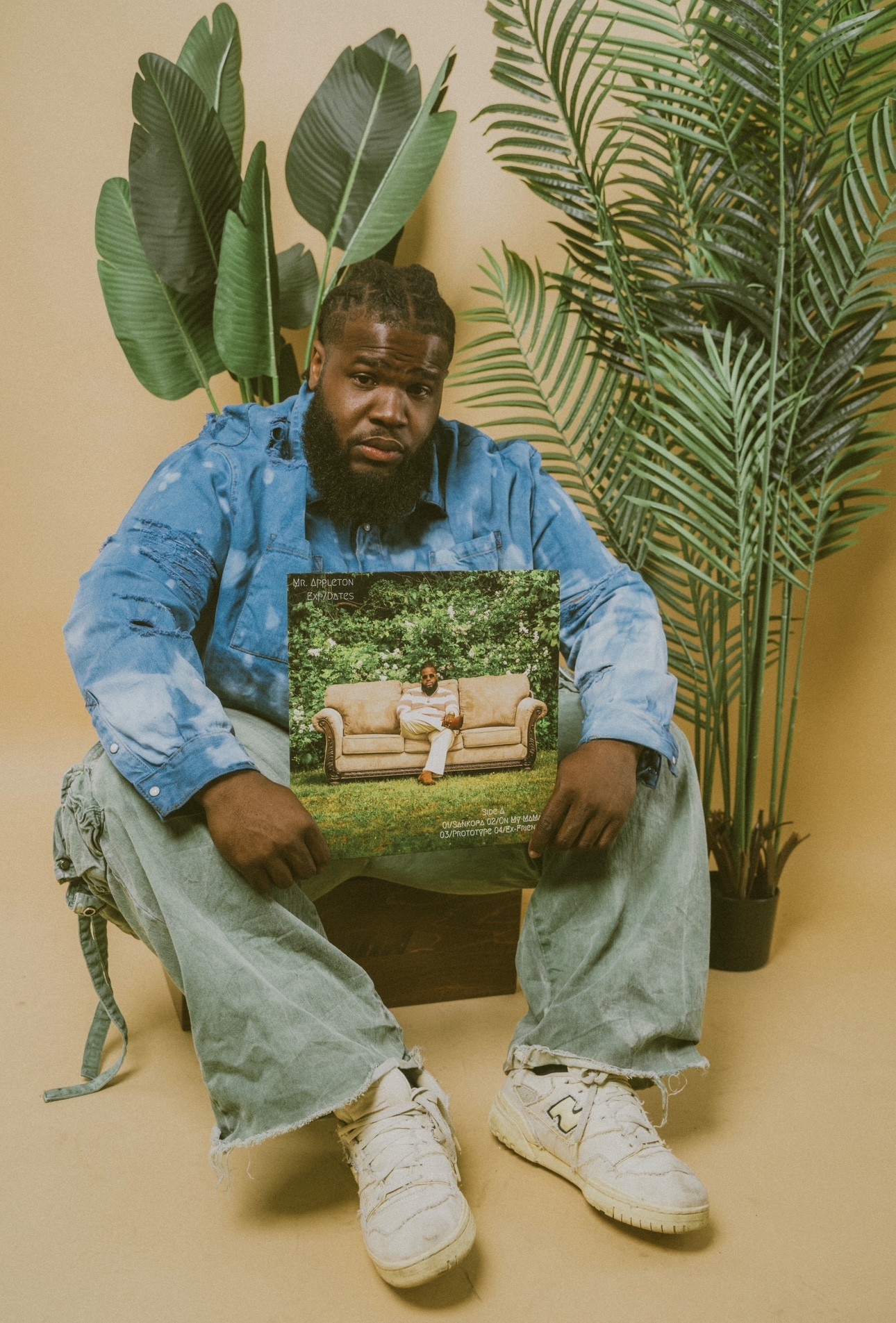 Man sitting on a chair holding a picture of a sofa outdoors, surrounded by large green plants, yellow background.