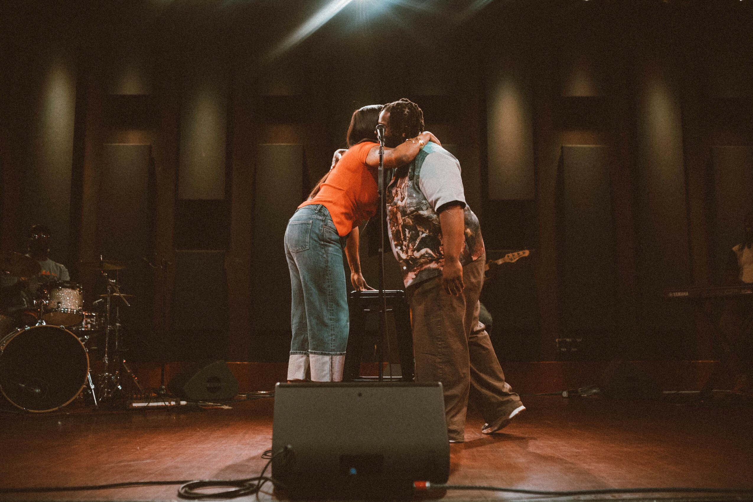 Two people embrace on stage with musical instruments in the background, dim lighting, and a dark backdrop.