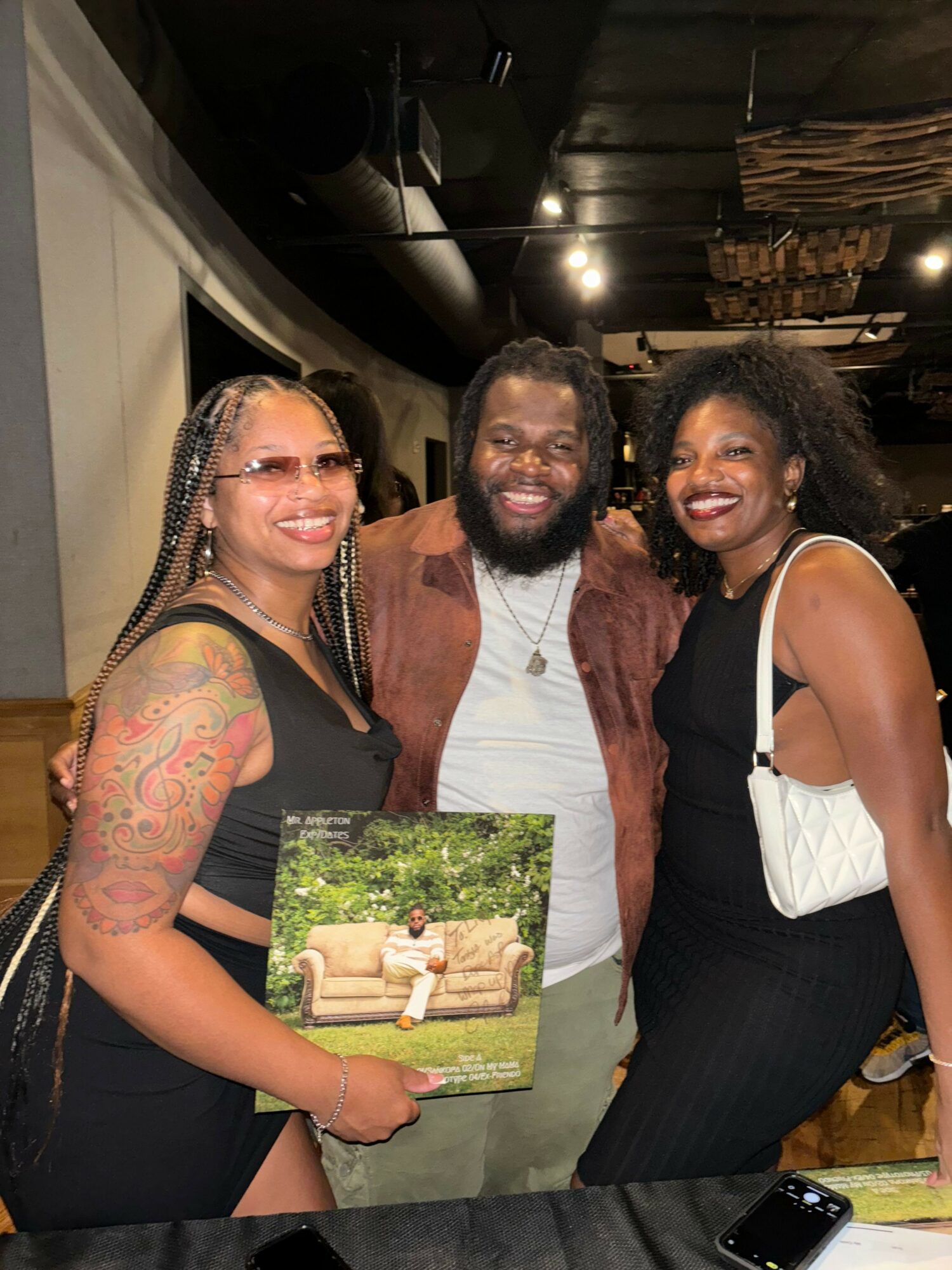 Three people smiling and posing together indoors, one with tattoos, one with curly hair, and one with glasses, holding a photo album.