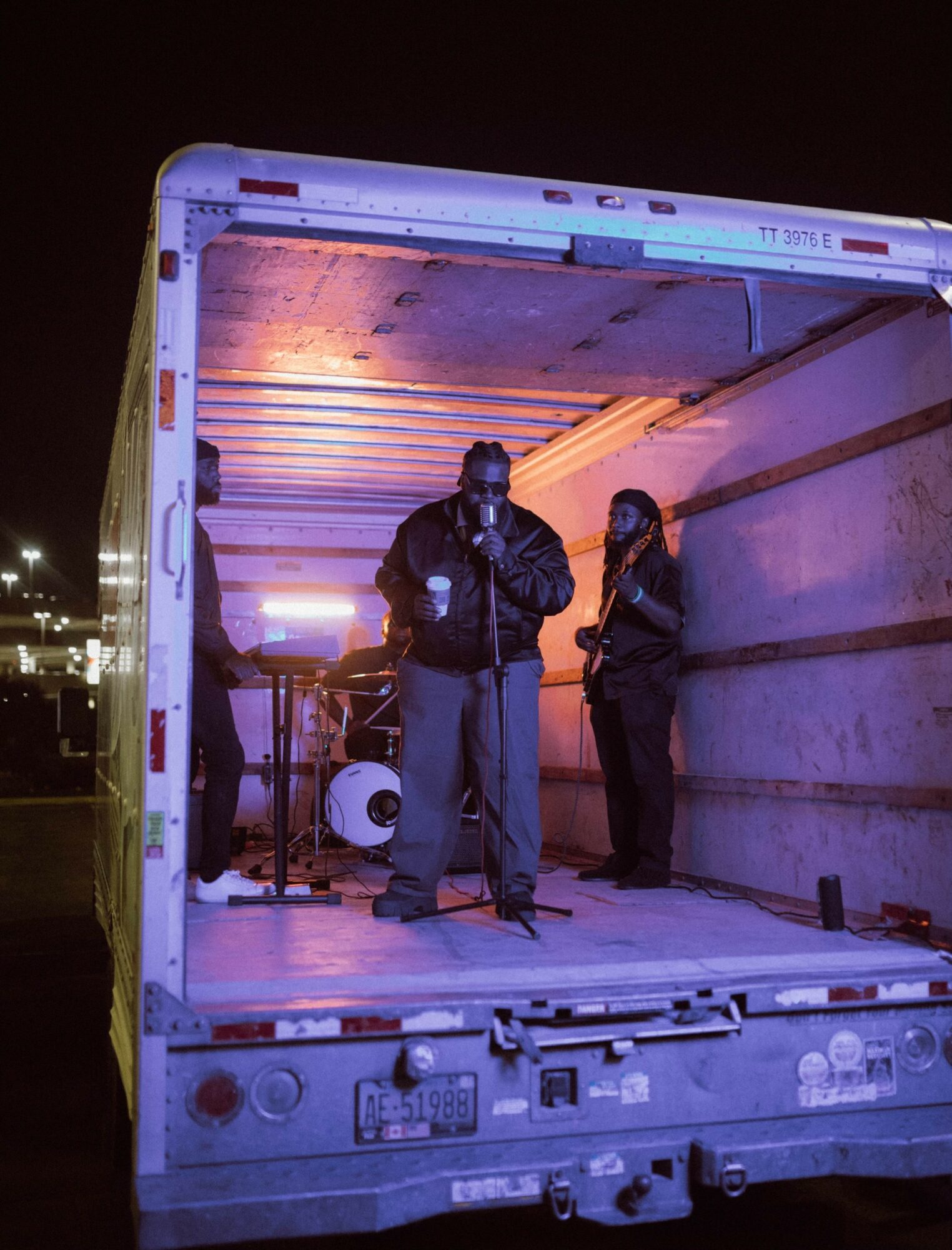 Three people stand inside an open truck at night, with one person speaking into a microphone, illuminated by interior lights.