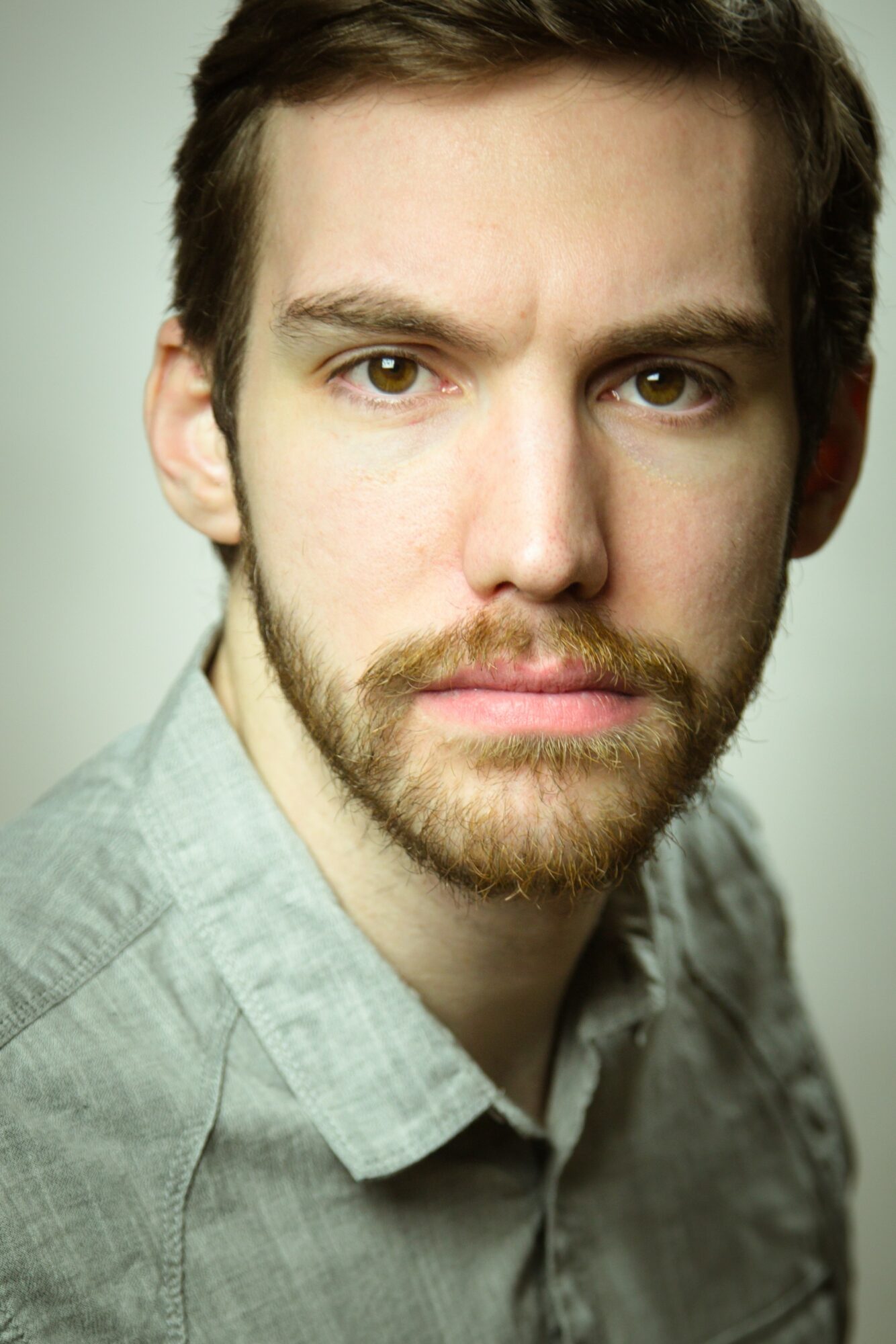 Close-up portrait of a young man with brown hair and beard, wearing a light gray shirt, looking at the camera.