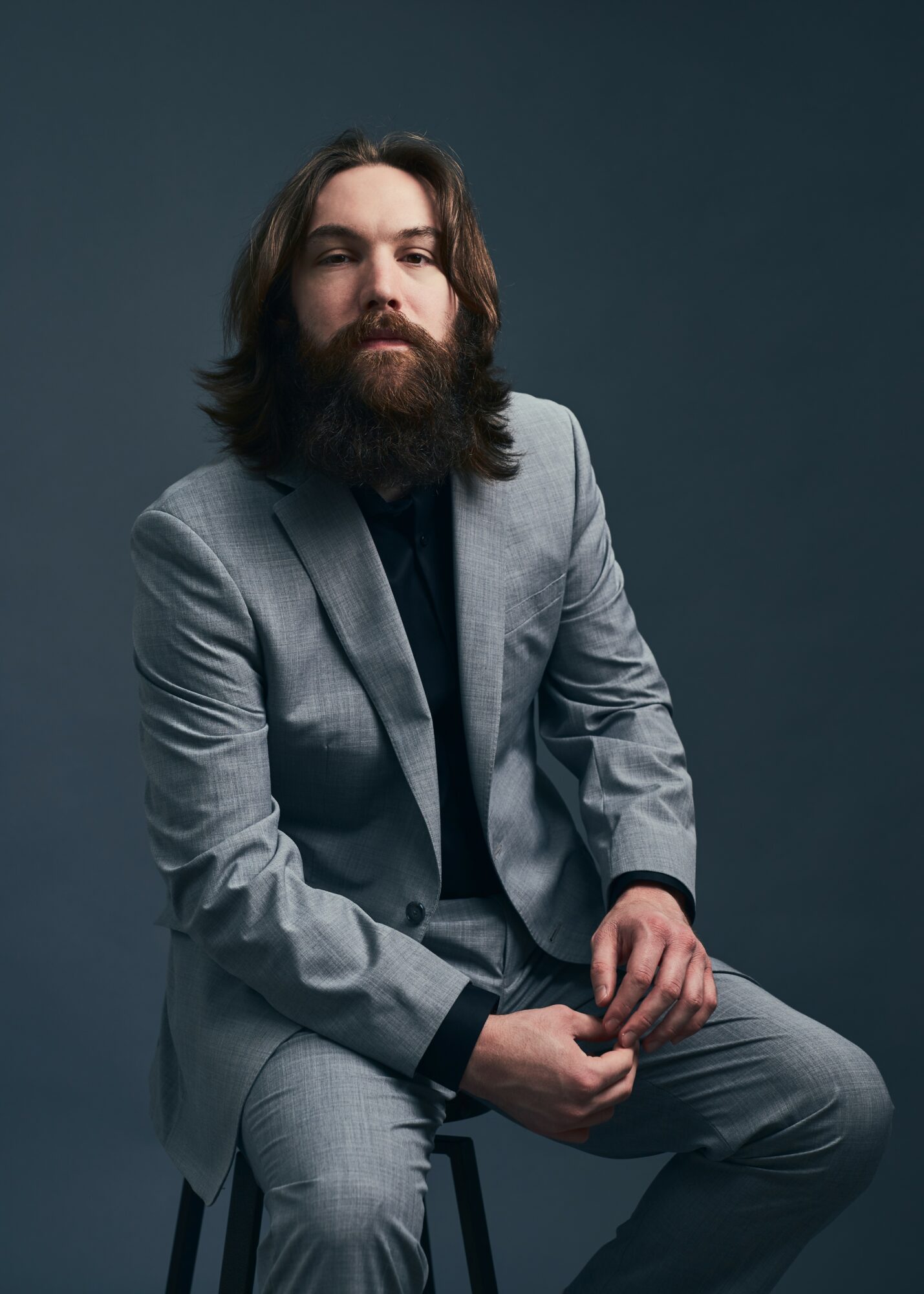 Man with long hair and beard in a gray suit sitting against a dark background.