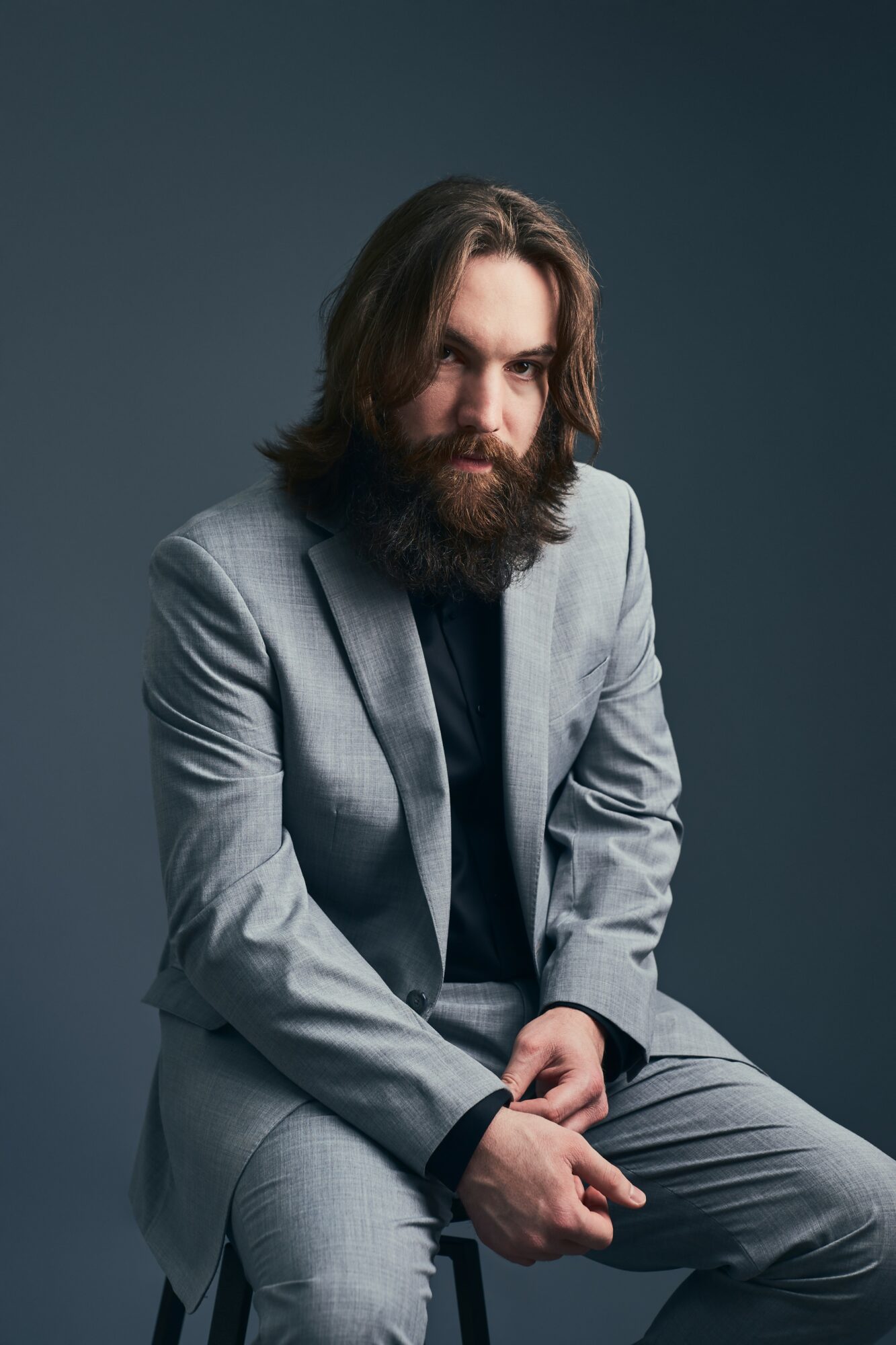 Man with long hair and beard in a gray suit sitting against a dark background.