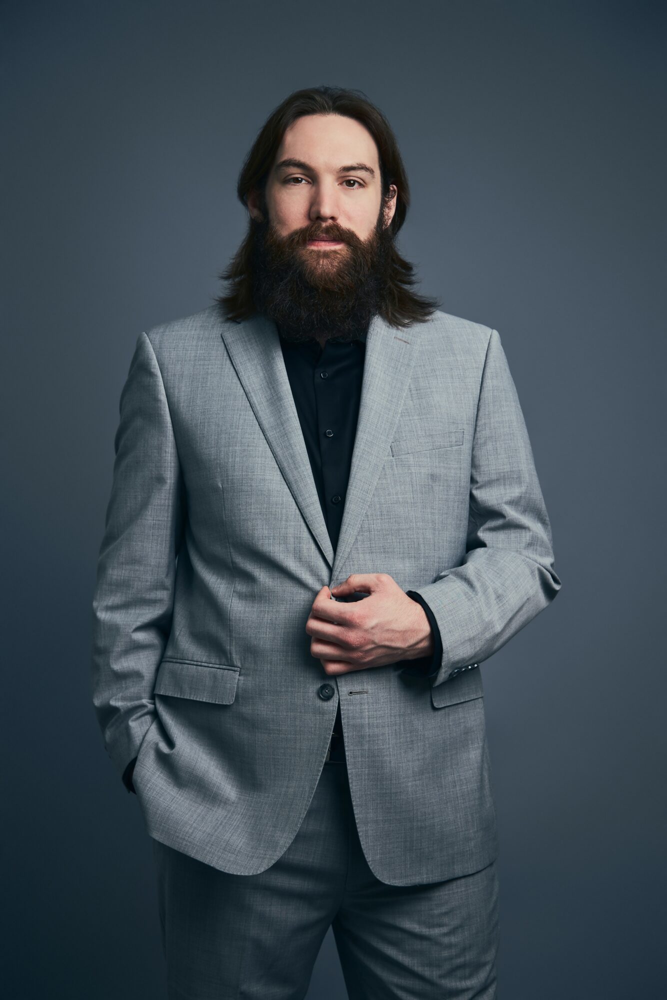 Man with long hair and beard wearing a light gray suit and black shirt, standing against a dark background.