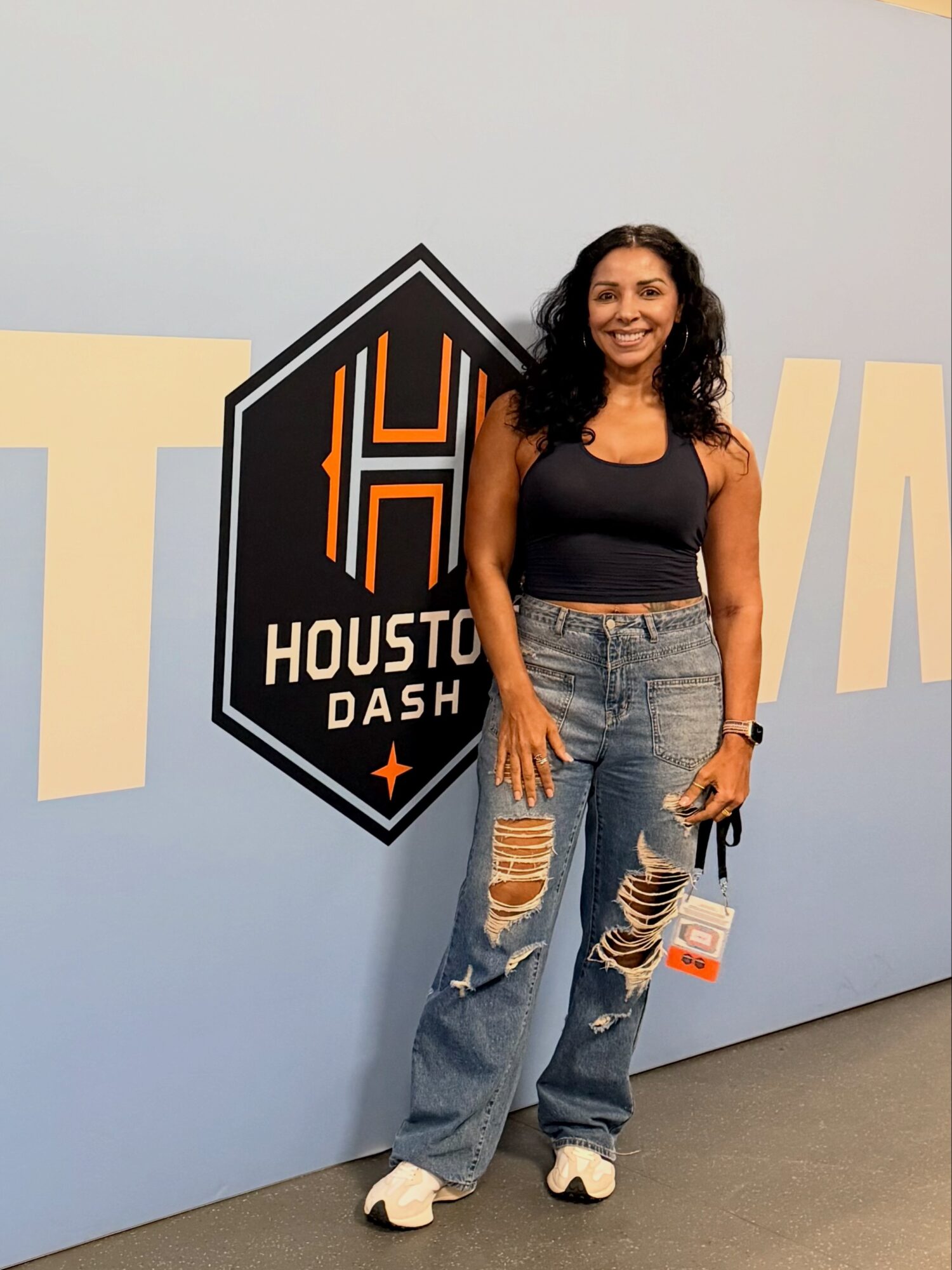 Woman with curly hair smiling, wearing a black tank top and ripped jeans, standing in front of a Houston Dash logo.