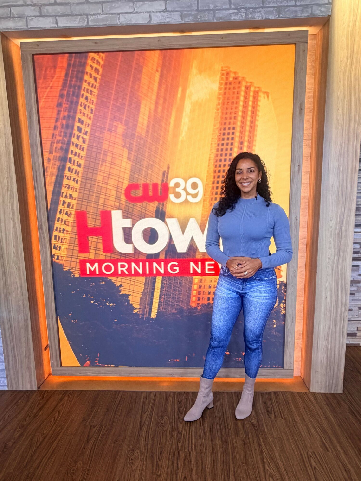 Woman standing next to a large TV screen displaying a morning news show with cityscape background.