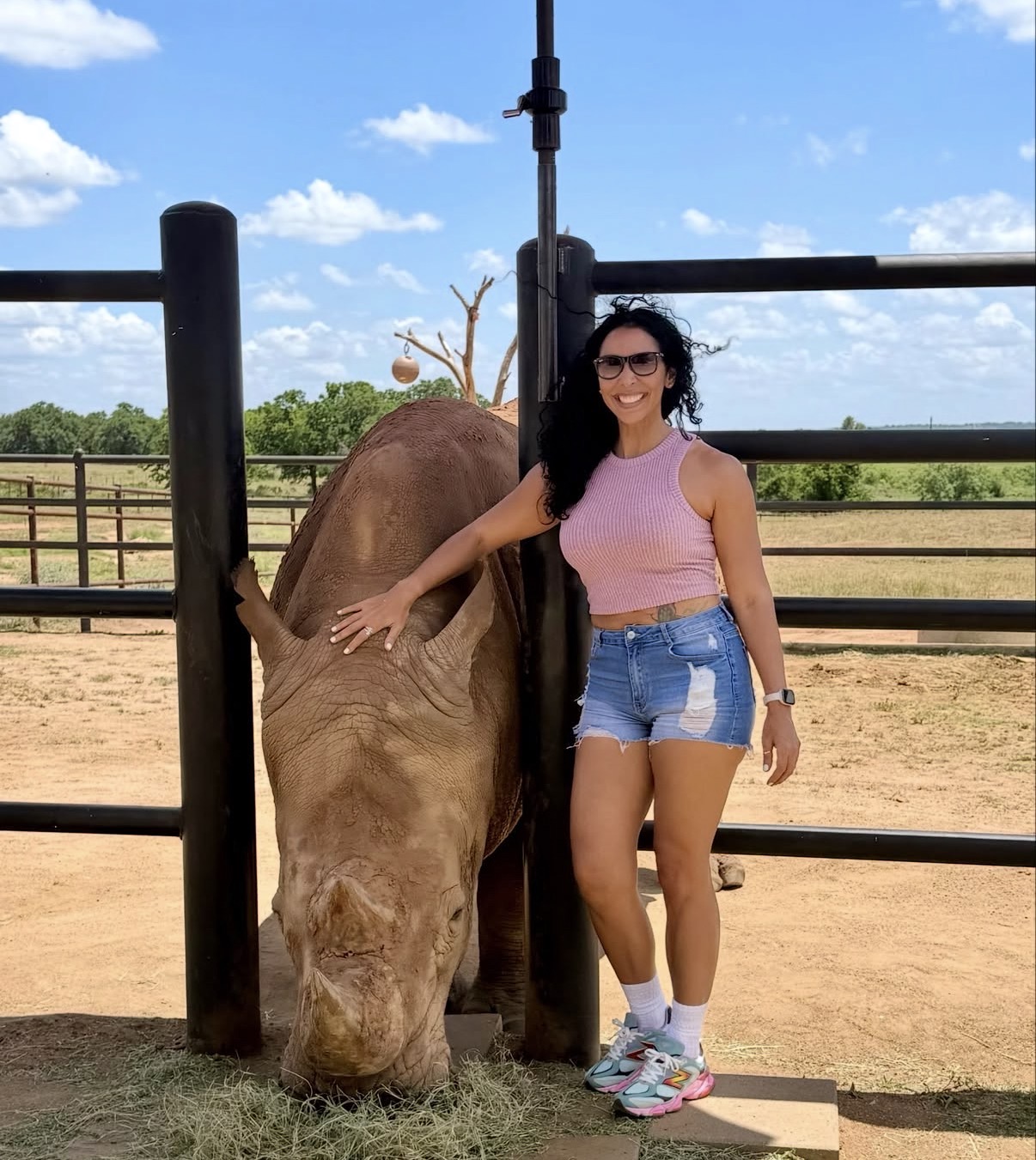 Woman in pink top and denim shorts standing next to a grazing horse in a fenced outdoor area under blue sky.
