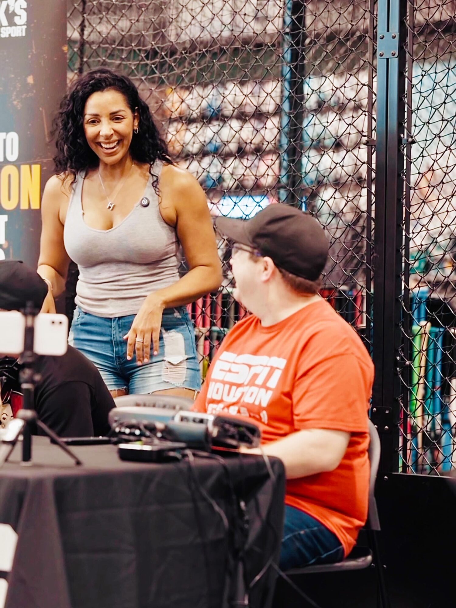Woman with curly hair smiling, wearing a gray tank top, standing outdoors near a chain-link fence, talking to a seated man in a red shirt and cap.