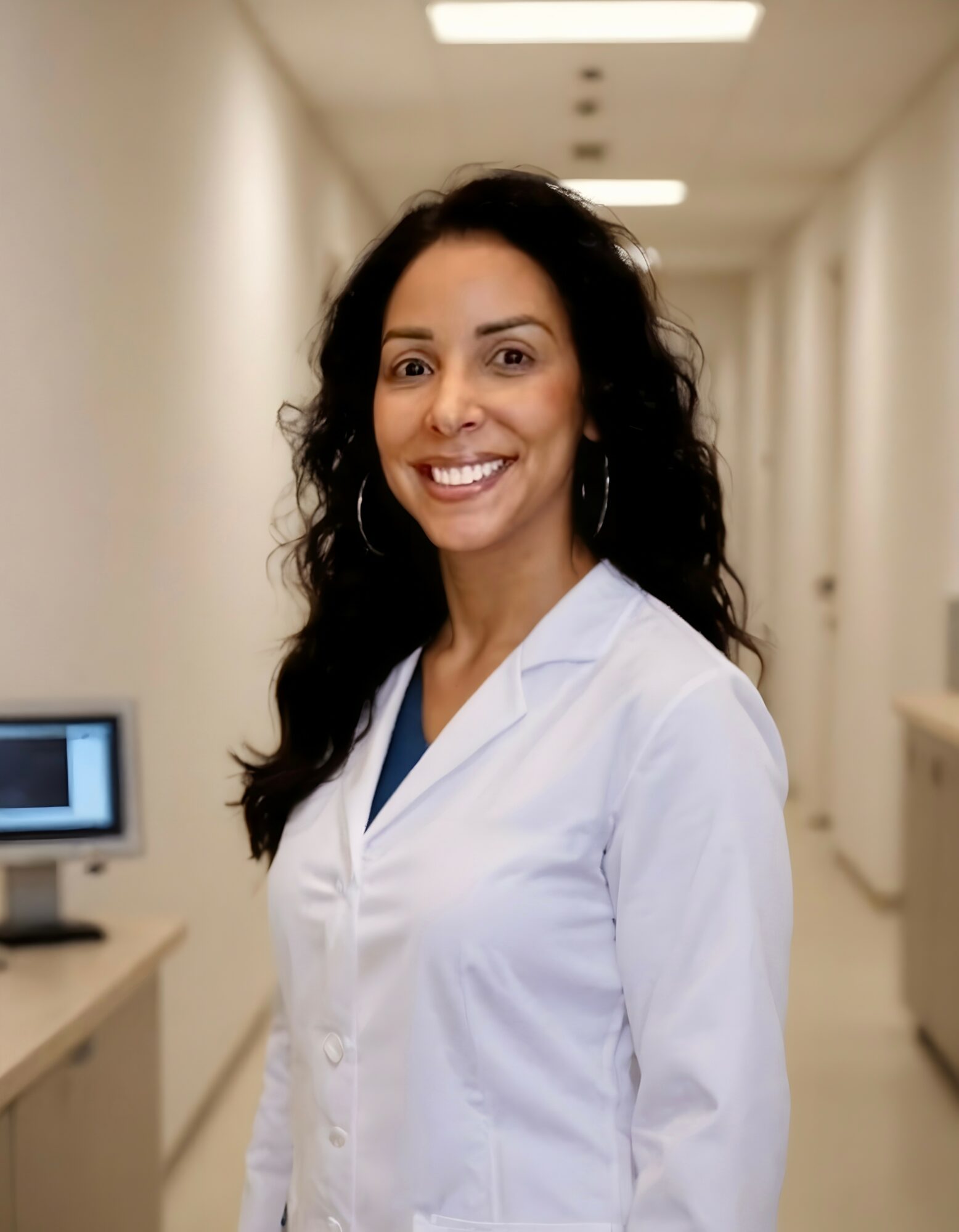 Smiling woman in white medical coat standing in a hallway with medical equipment, facing forward.