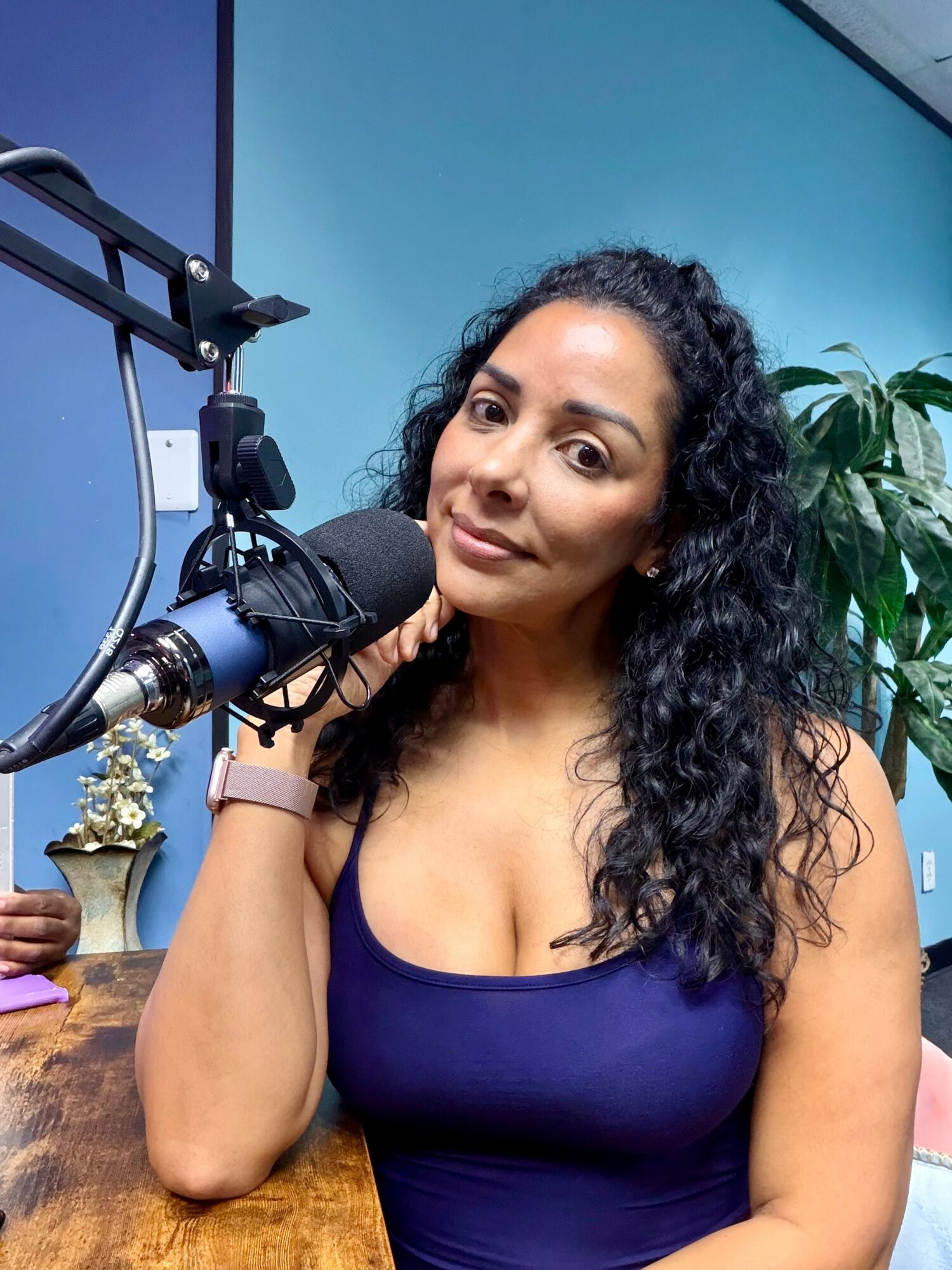 Woman with curly hair sitting at a desk with a microphone, in a room with blue walls and a plant.