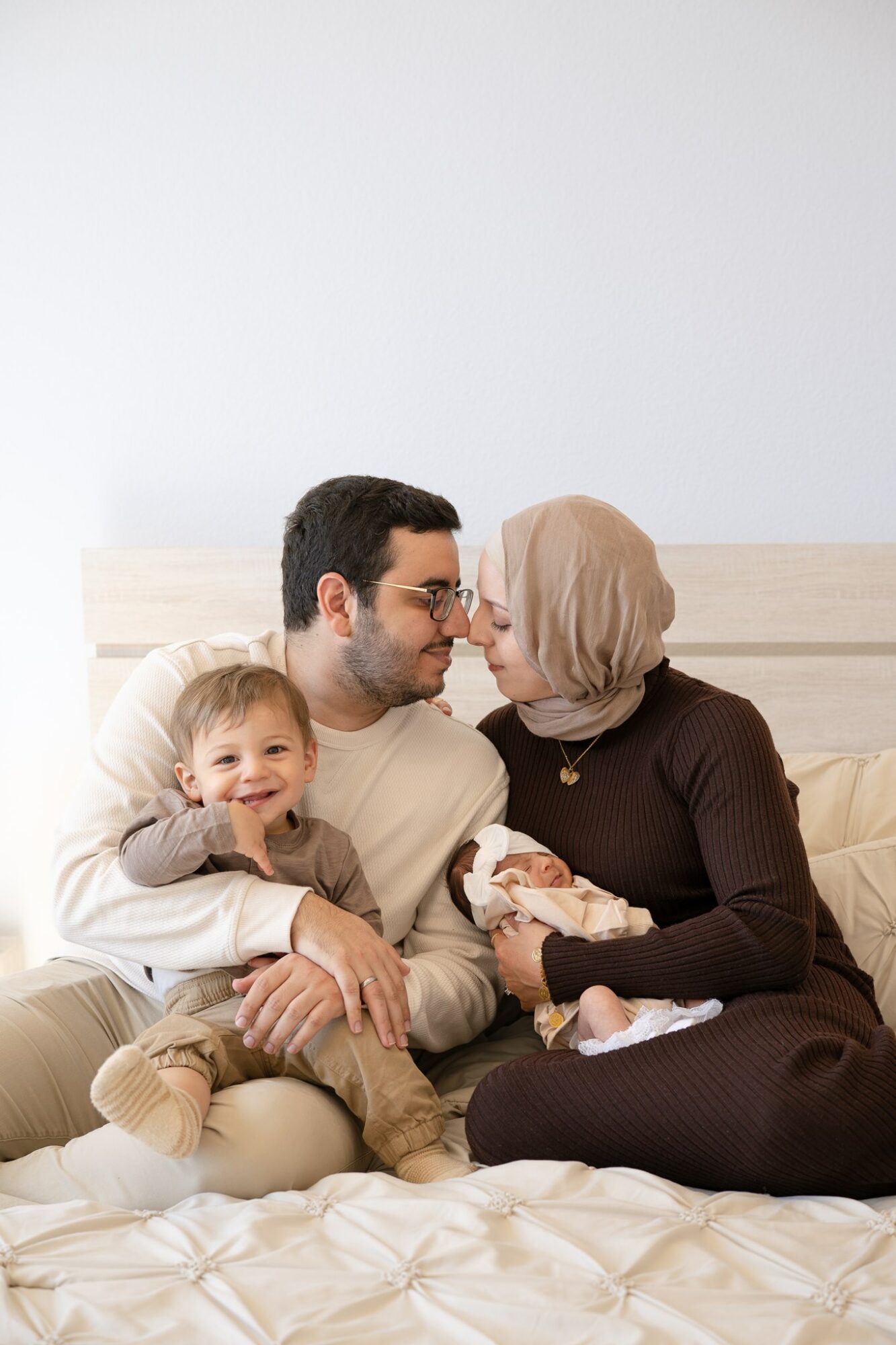 Family sitting on bed, woman holding baby, young girl smiling, man and woman looking at each other, light background