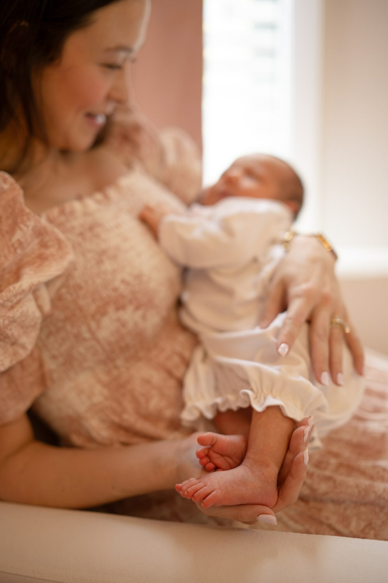 Woman holding a baby, smiling, indoors near a window, woman wearing a pink lace top, baby in white dress with ruffled hem, barefoot.