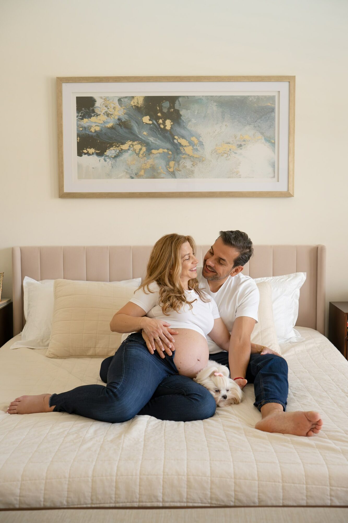 A couple sits on a bed with a dog, smiling and embracing, in a bedroom with a framed abstract painting above the headboard.