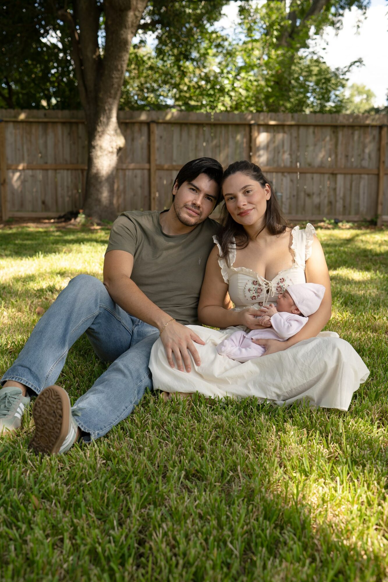 Family sitting on grass in backyard, woman holding baby, man leaning close, wooden fence and trees in background.