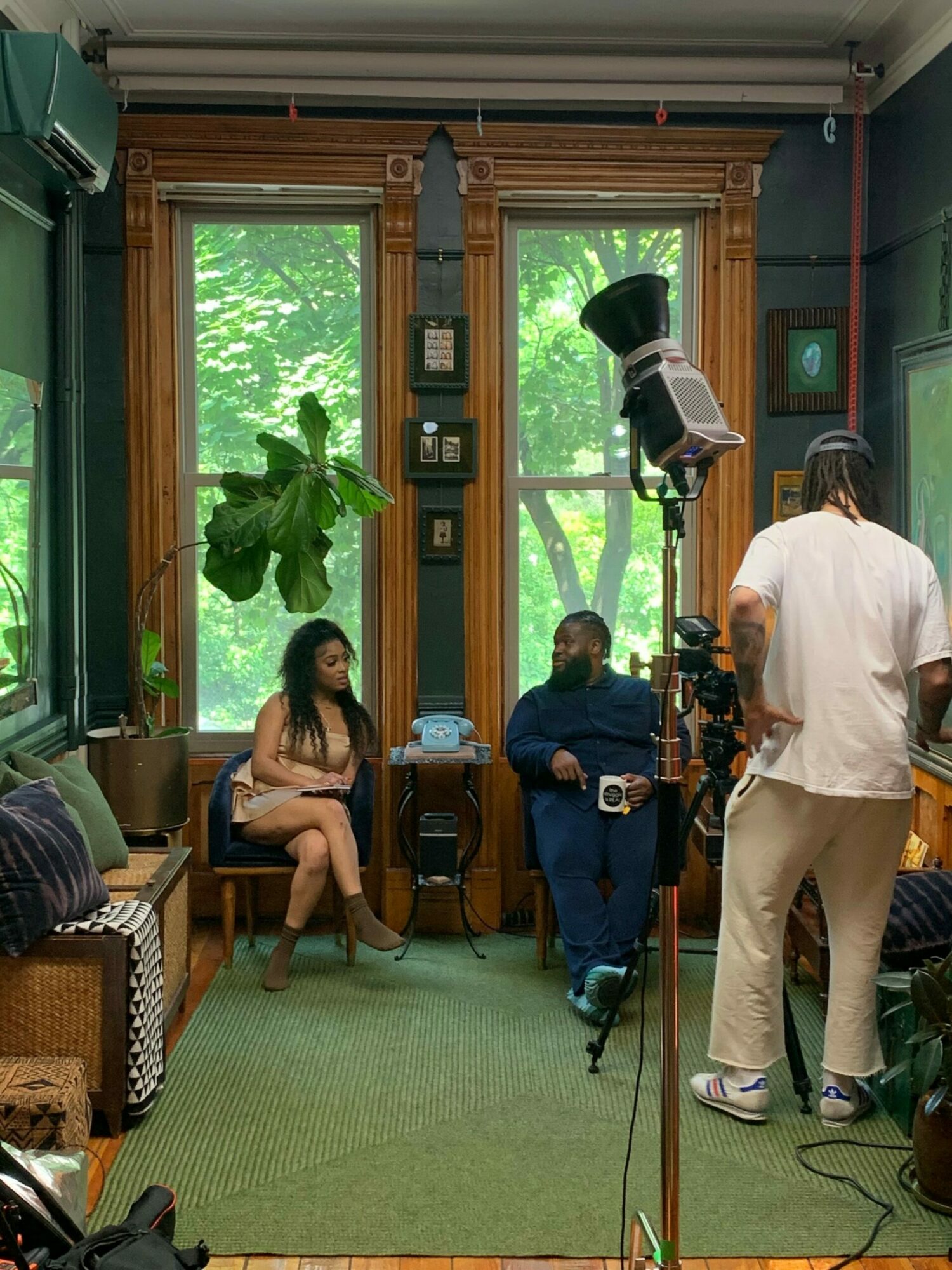 Two people in a room with large windows and green trees outside, one sitting on a bench and the other standing, with camera equipment around.