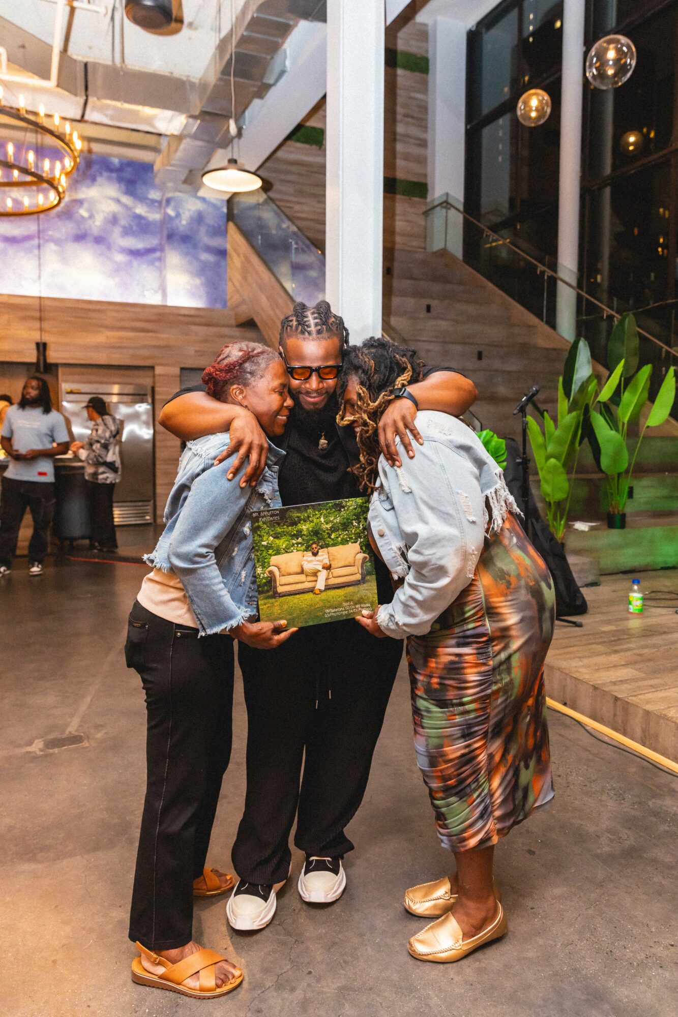 Three people embrace and look at a gift box in a modern indoor space with plants and large windows.