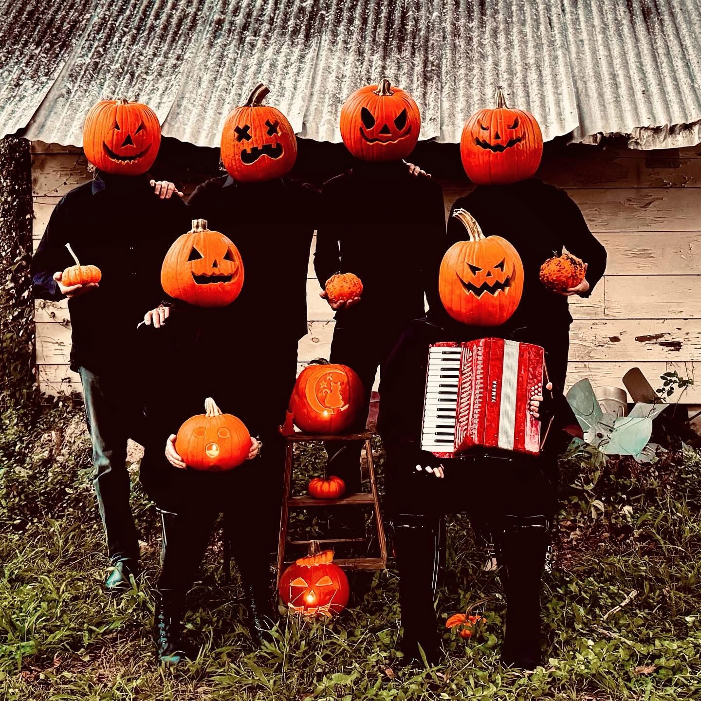 Group of people with pumpkin masks, some holding pumpkins, one with an accordion, standing outdoors in front of a rustic building.