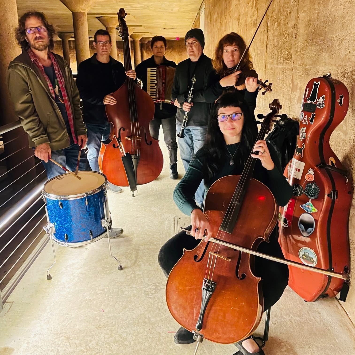 Group of six musicians with instruments, including a cello, violin, double bass, and drums, in a hallway with warm lighting.