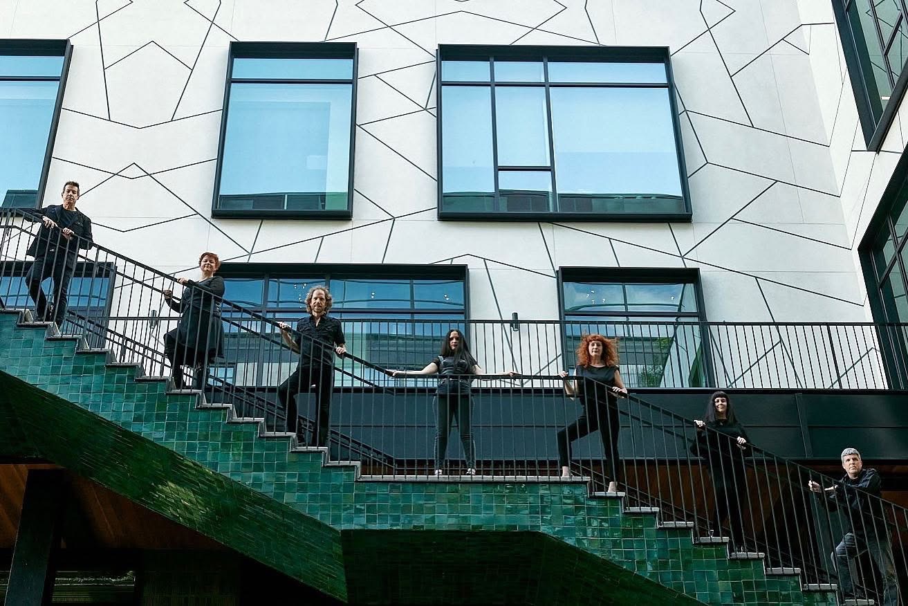 Six people standing on a green staircase with a modern building in the background, windows reflecting the sky.