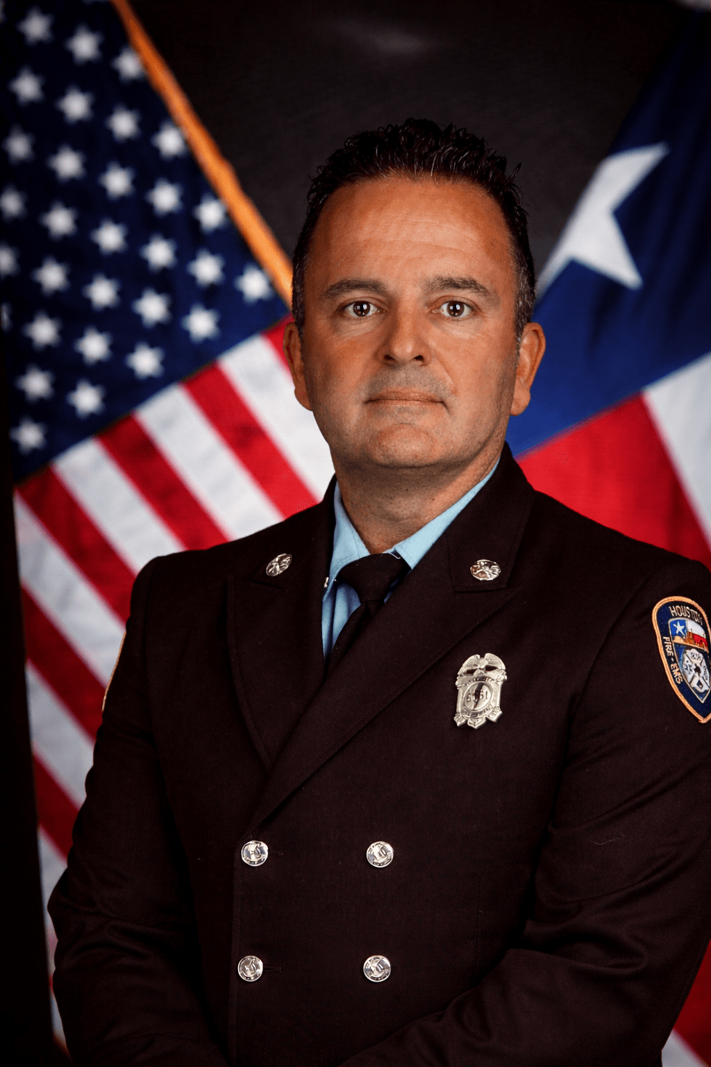 Firefighter in uniform standing in front of American and Texas flags, looking at the camera, serious expression.