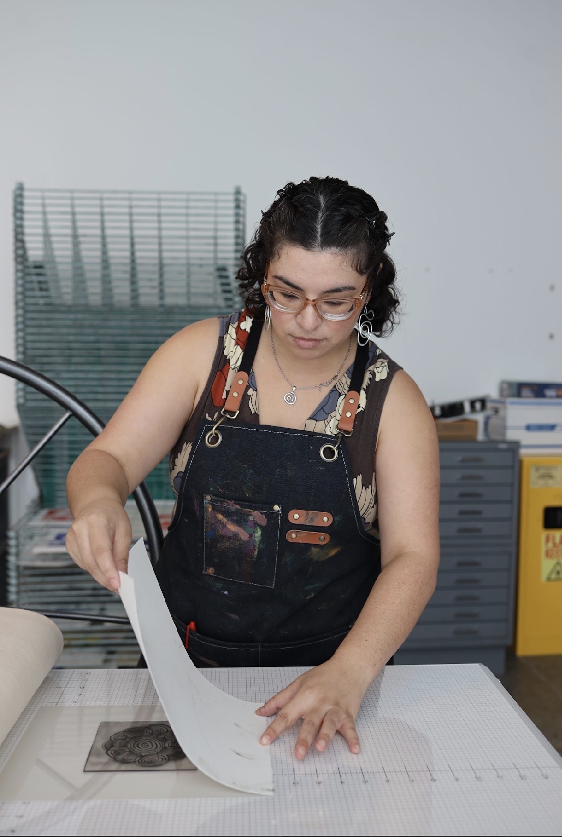 Woman with glasses and curly hair wearing overalls, working with paper on a table in a room with storage items.
