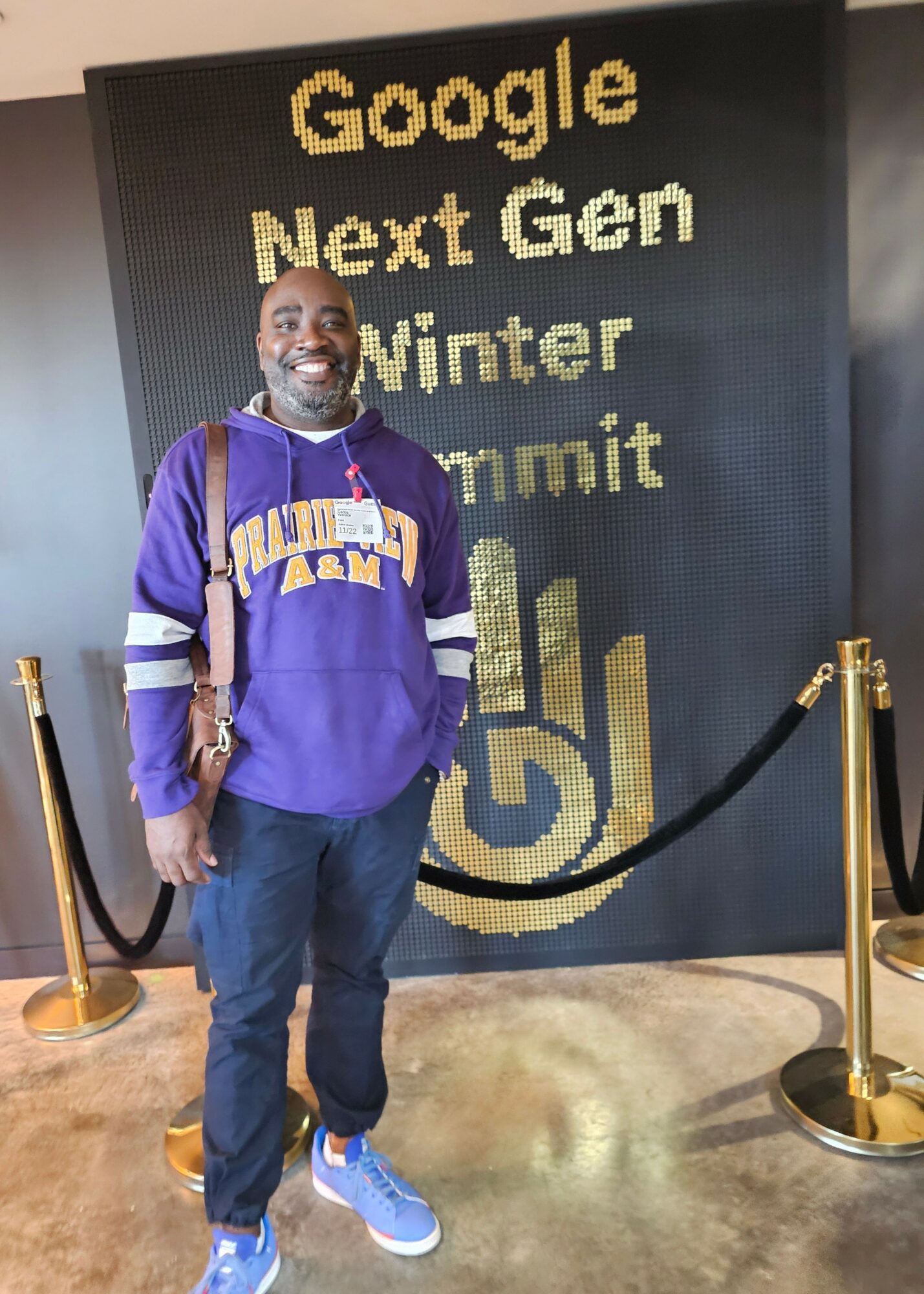 Man smiling in front of a large digital display at Google Next Gen Winter Summit, wearing a purple hoodie and jeans.