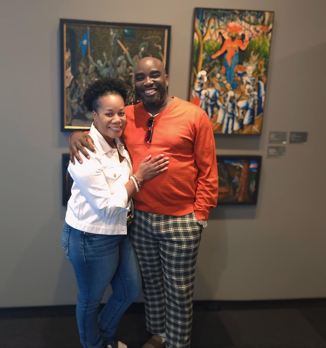 Two people smiling and posing together in an art gallery, with colorful paintings on the wall behind them.
