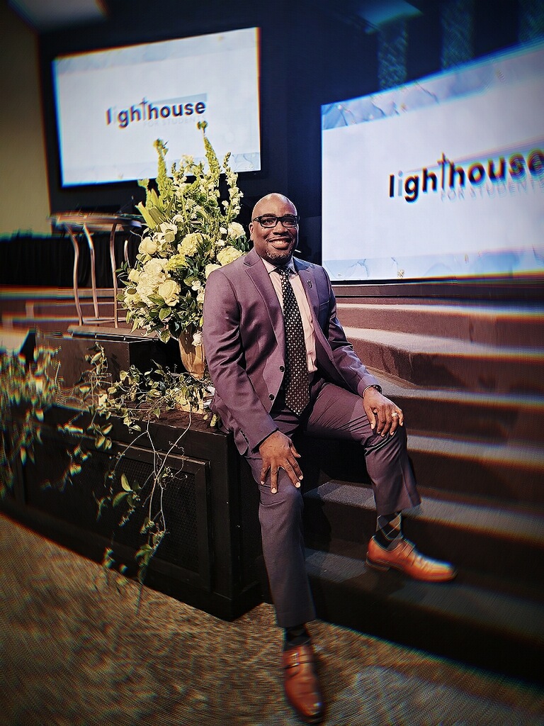 Man in suit sitting on stage with large flower arrangement and screens displaying 'lighthouse' behind him.