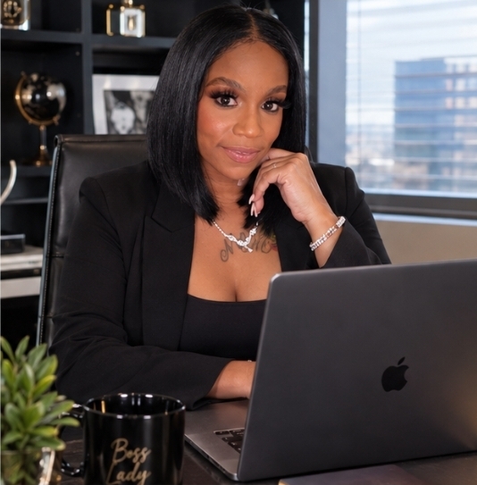 Woman with black hair in a black blazer sitting at a desk with a laptop, window view in background.