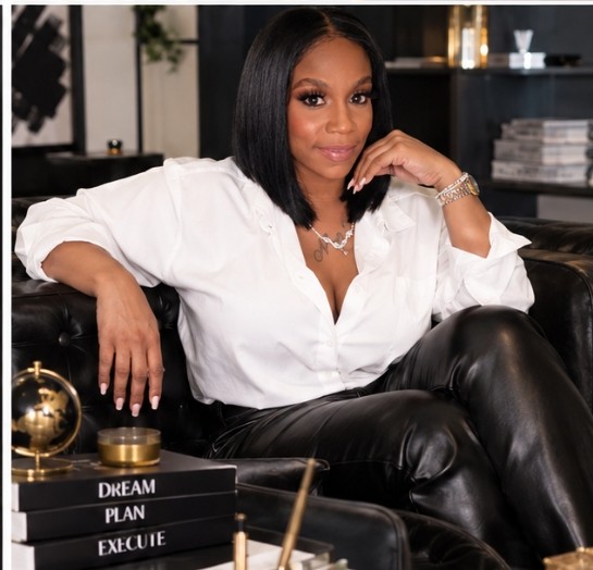Woman with shoulder-length black hair, wearing a white shirt and black leather pants, sitting on a black sofa in a modern room.