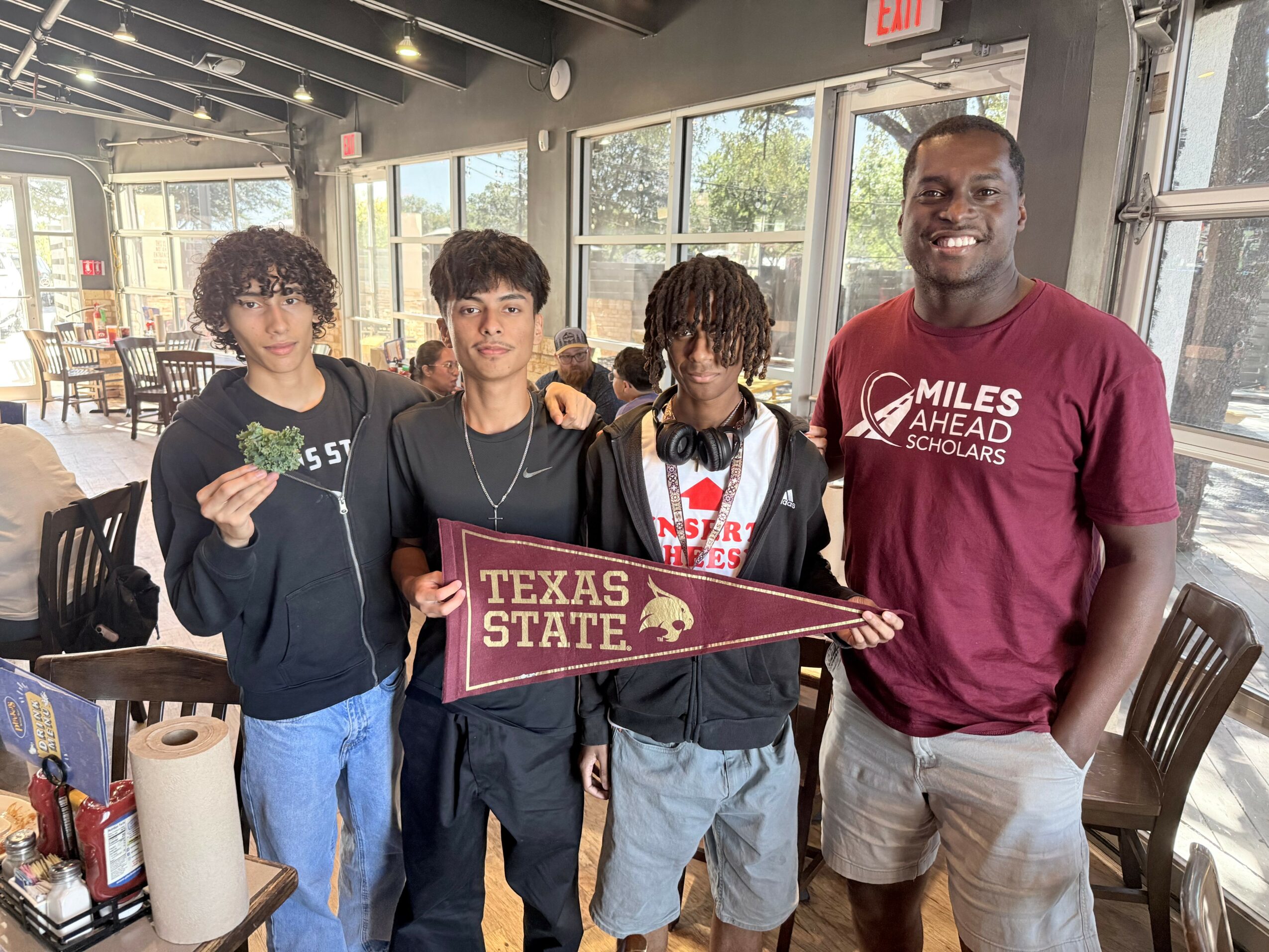 Four people standing indoors, holding a Texas State University flag, smiling, with large windows and tables in the background.