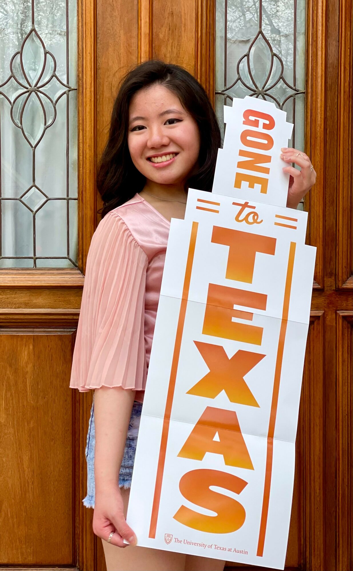 Young woman smiling holding a large Texas-themed sign in front of wooden and glass door background.