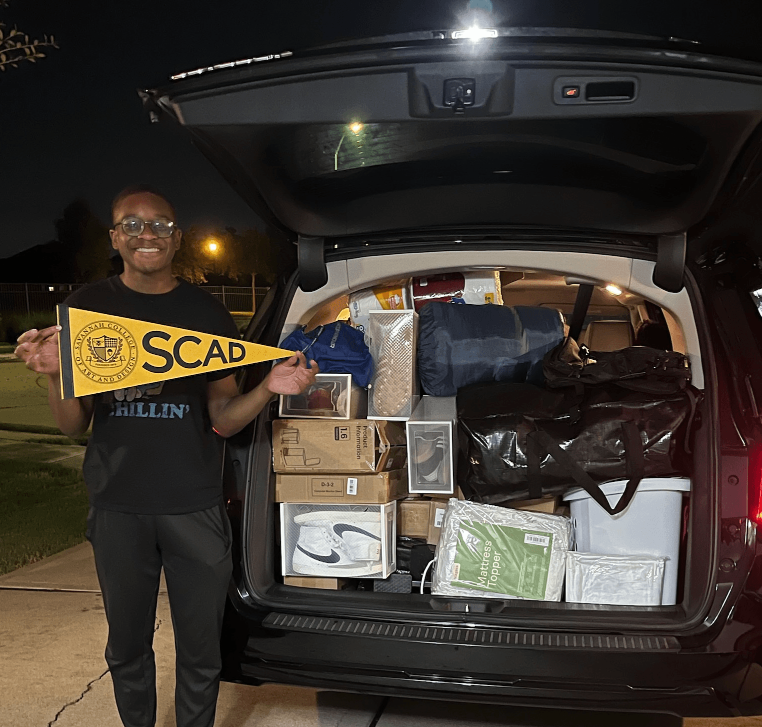 Person standing next to an open vehicle trunk filled with boxes and bags, holding a yellow SCAD pennant, nighttime setting.