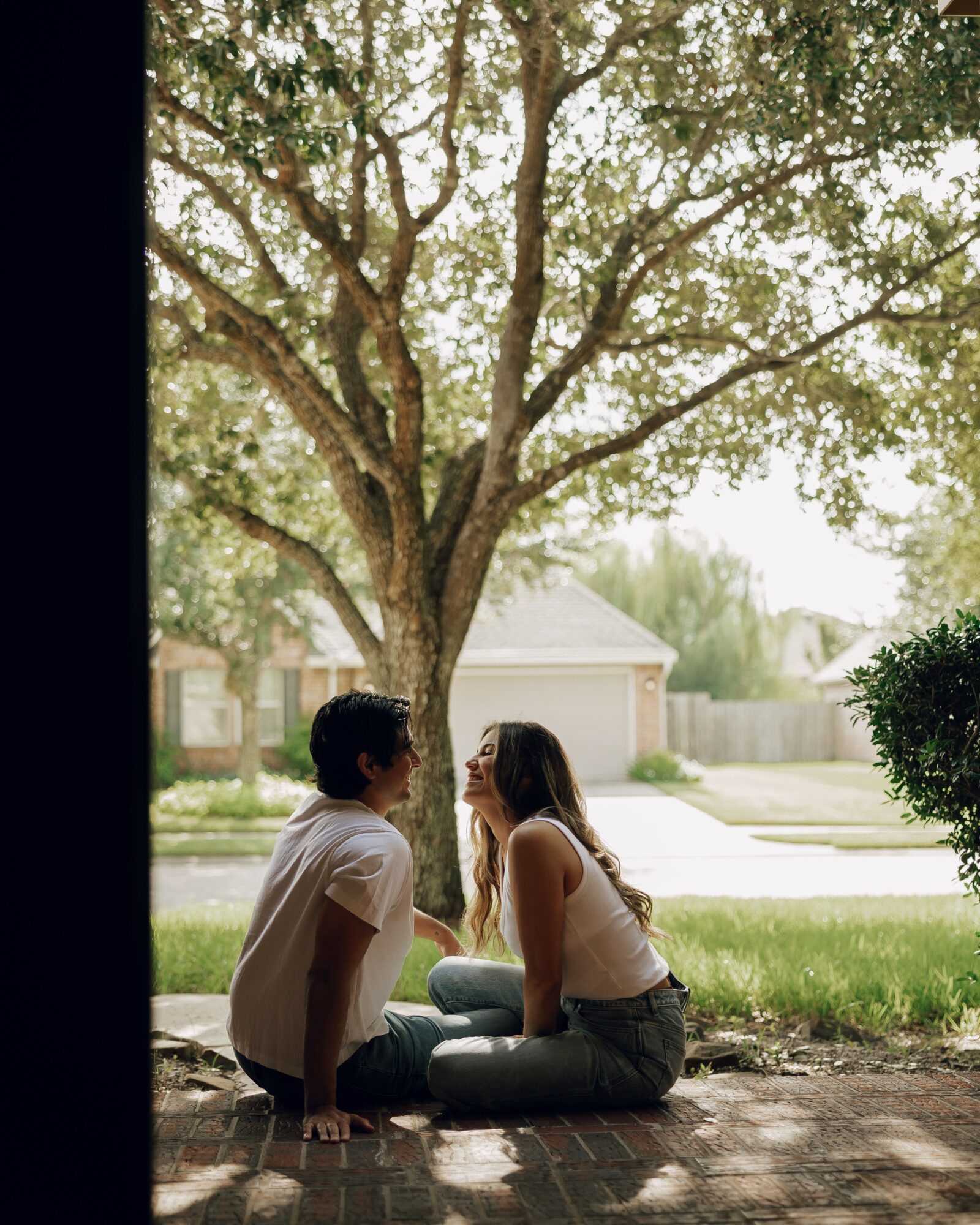 Two people sitting on a brick porch under a large tree, facing each other and talking, with a suburban street in background.