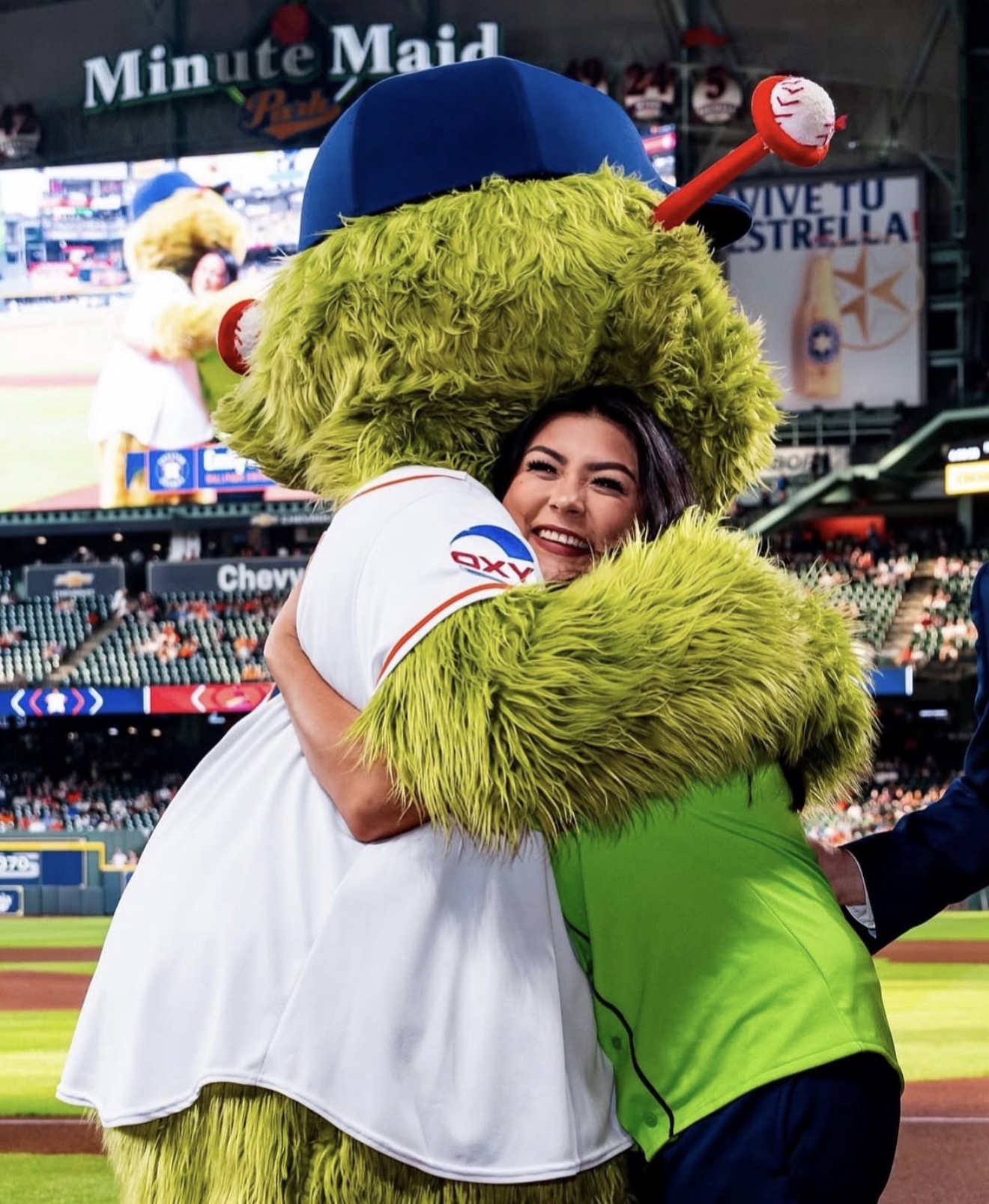 Person hugging a large green furry mascot in a baseball stadium, smiling, with fans and advertisements in background.