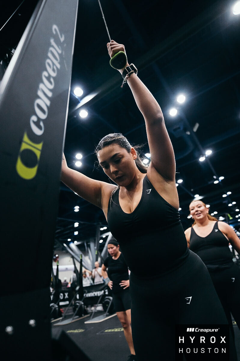Female athlete in black sportswear adjusts equipment at a gym, with other women in the background, bright overhead lights.