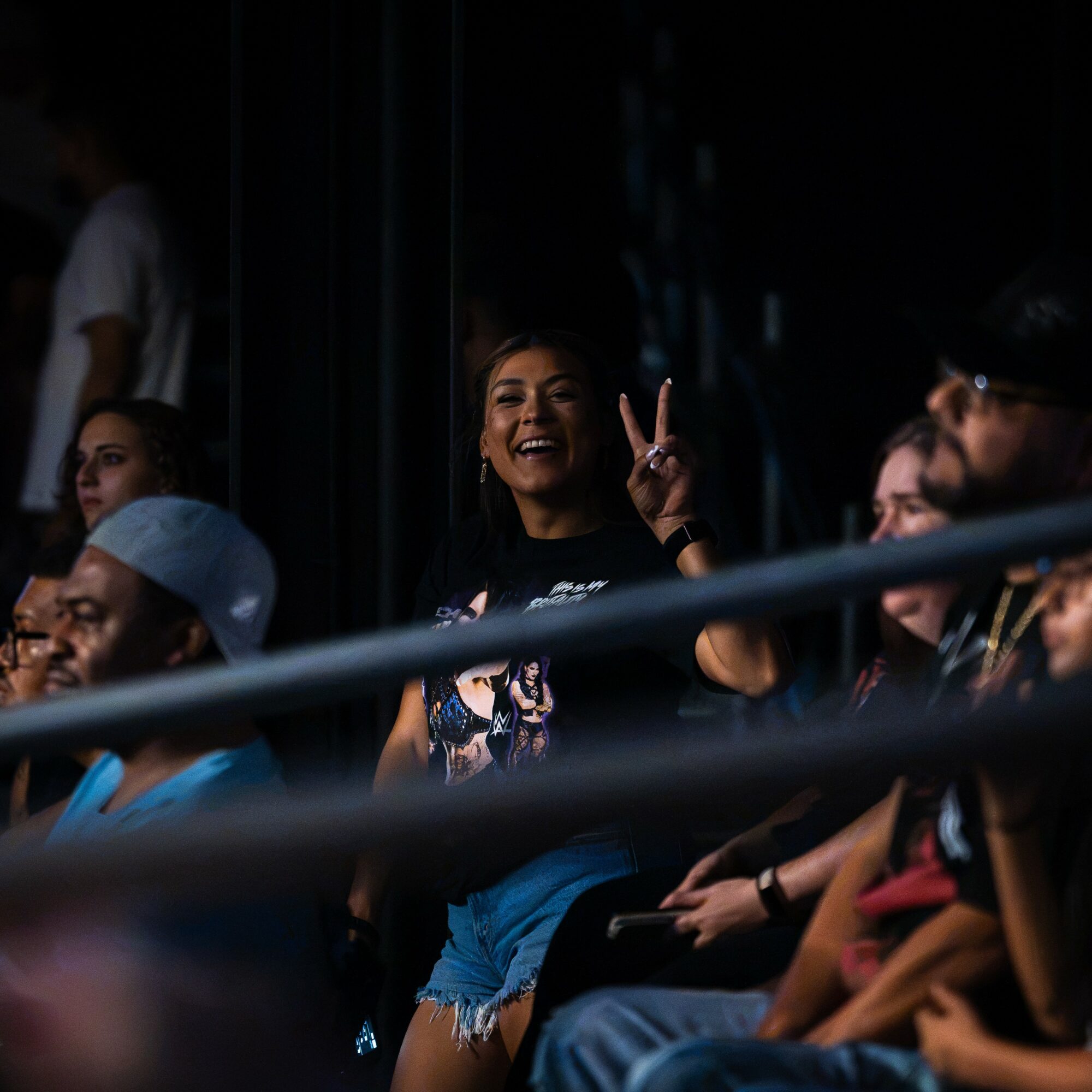 Group of people sitting in a dark venue, woman smiling and making a peace sign, others watching attentively.