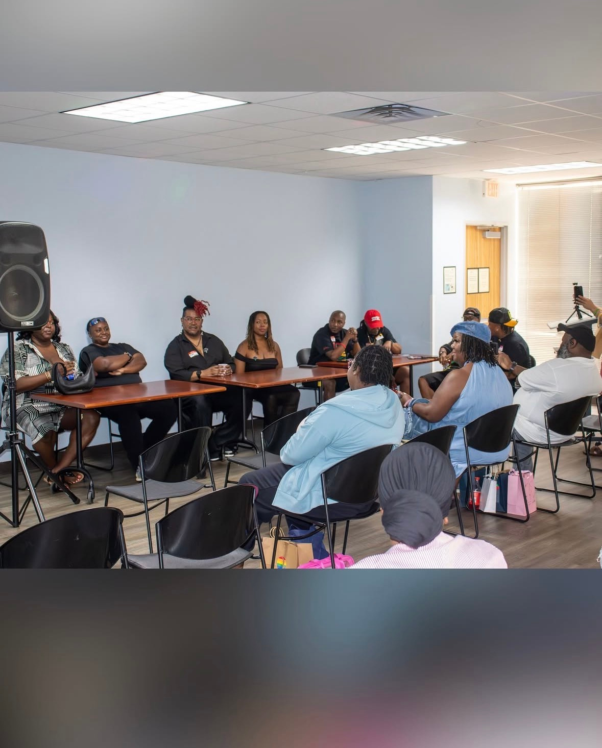 People seated around a table in a meeting room, some wearing hats, with a speaker and a door in the background.
