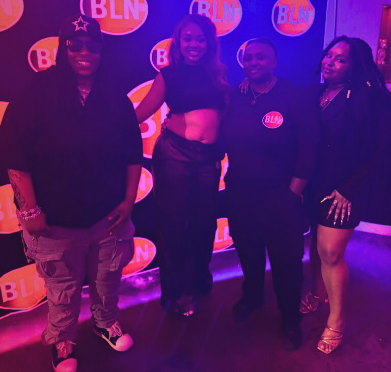 Four women standing together in front of a backdrop with circular logos, smiling and posing for the photo.