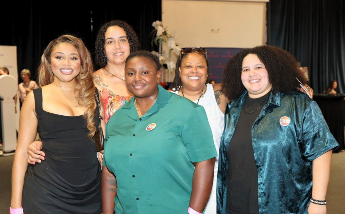 Five women standing together indoors, smiling, with a stage and black curtains in the background.