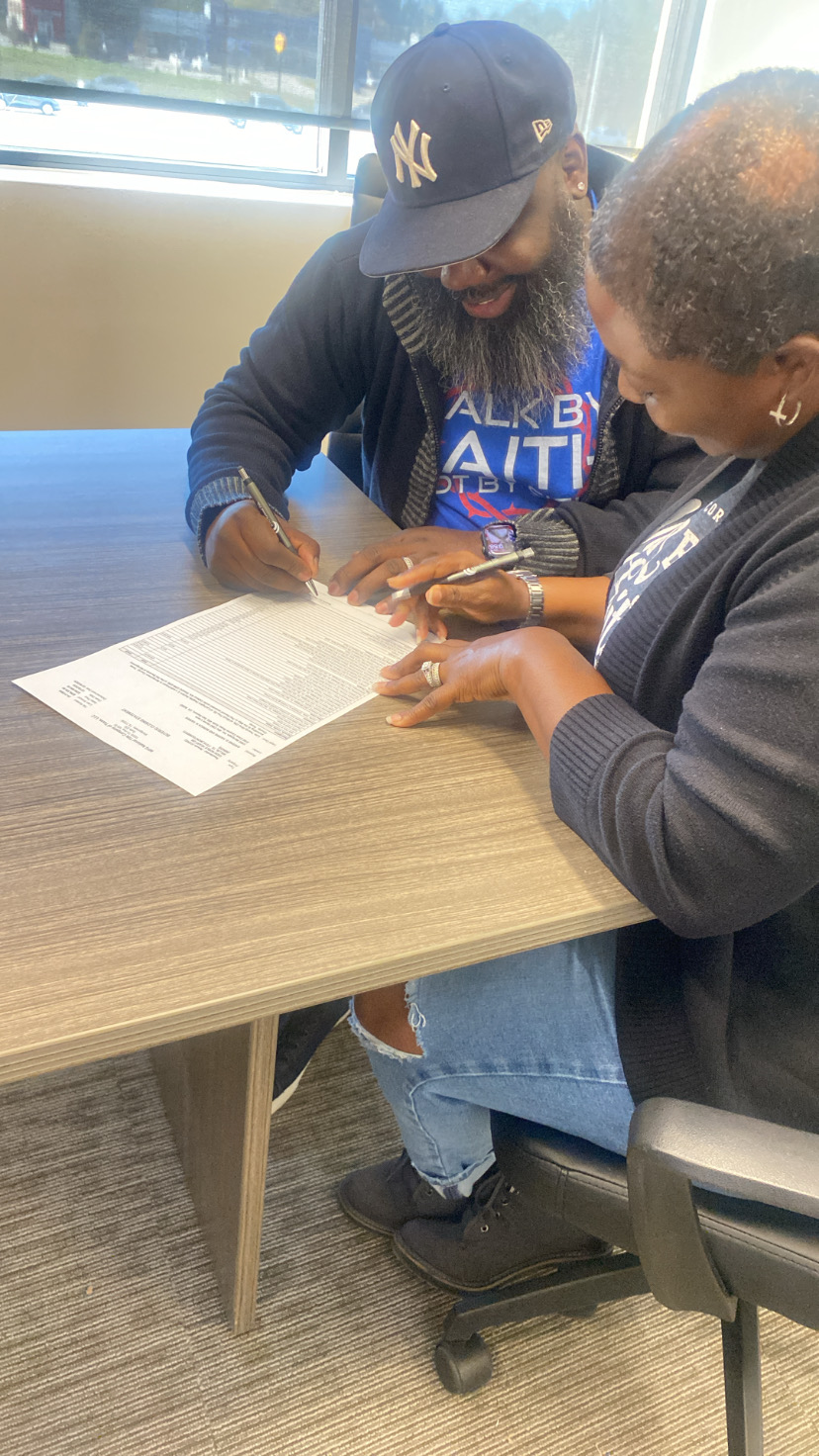 Two women sit at a table, signing a document, with a window in the background.