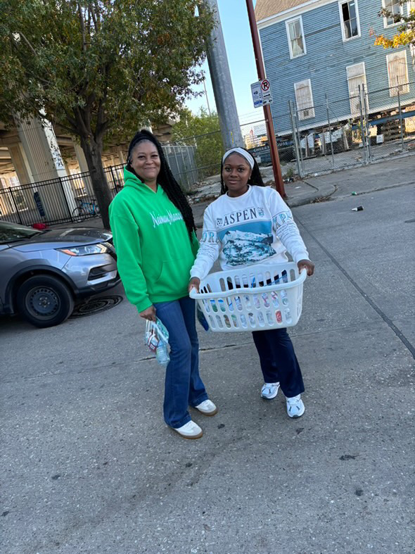 Two women standing outdoors, one holding a laundry basket, with trees, cars, and buildings in the background.