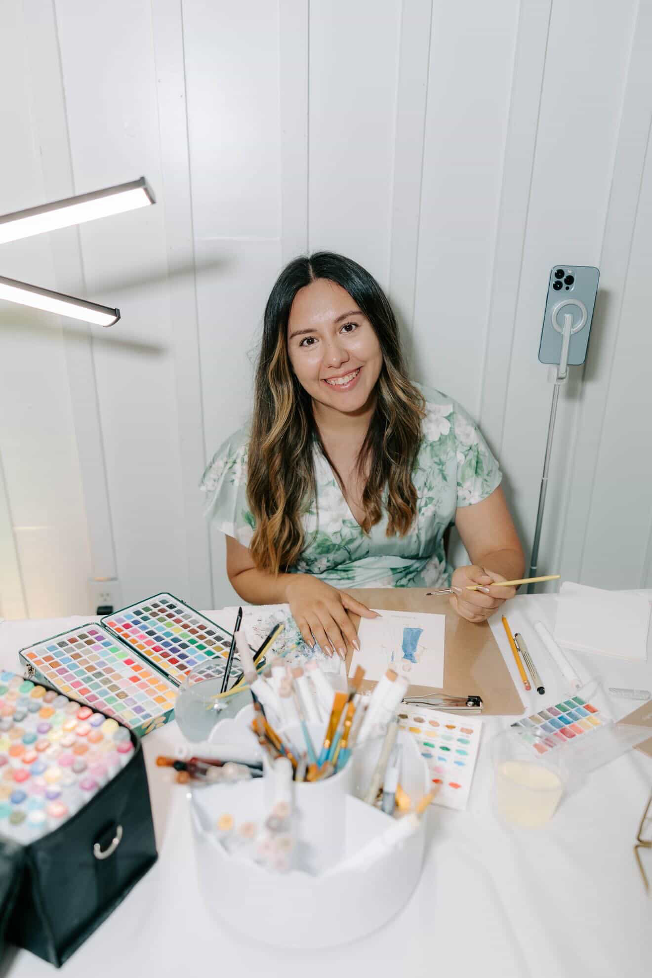 Woman sitting at a table with art supplies, smiling, white wall background, light-colored hair, floral top, various art materials on table