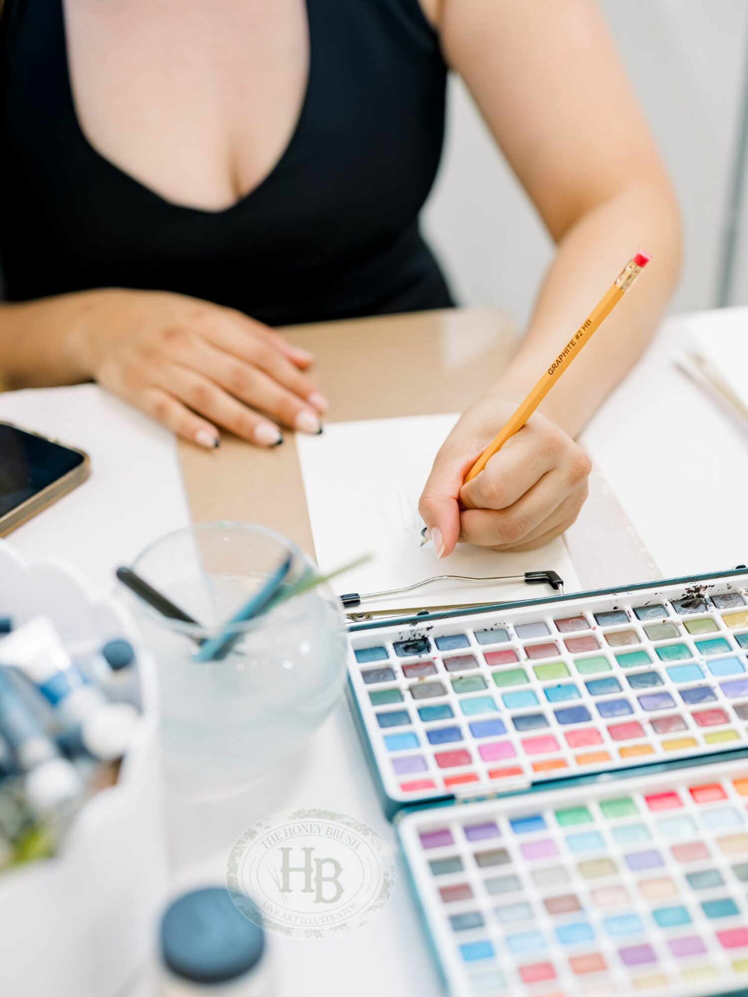 Person writing with a pencil at a desk with watercolor paints and paper, wearing a black top.