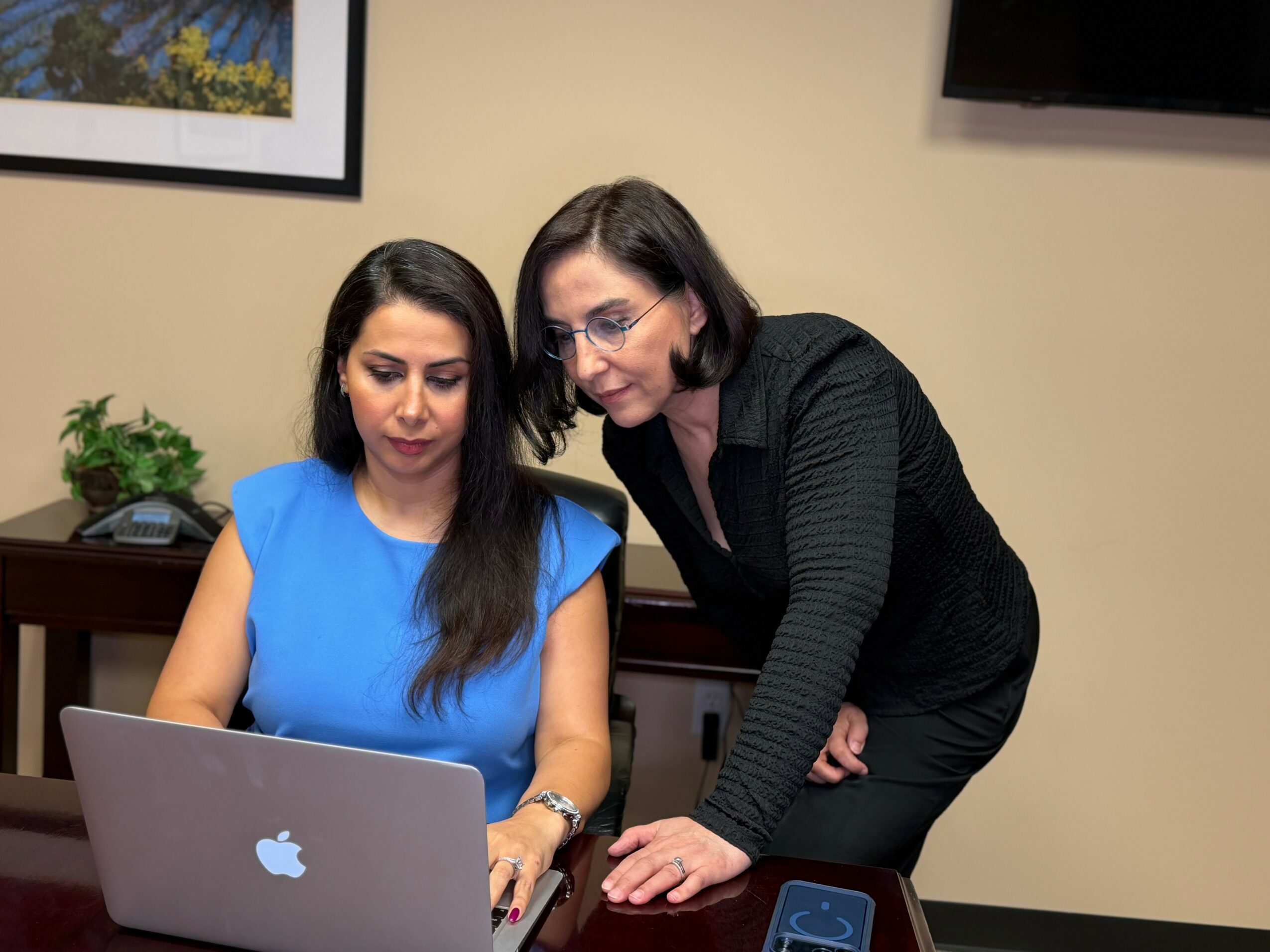 Two women look at a laptop screen in an office, one seated and one standing, with a plant and picture on the wall.